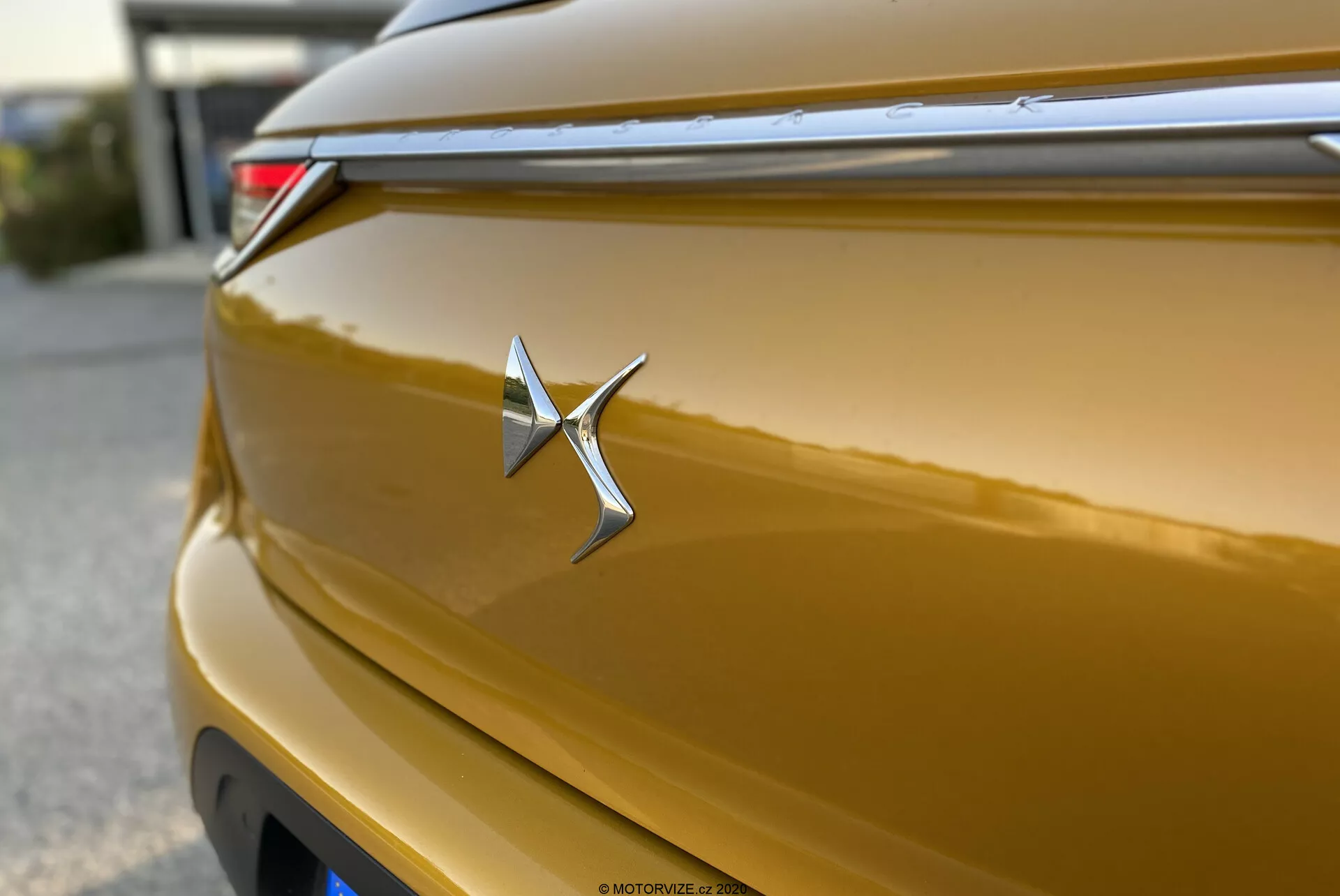 Primo piano di un badge DS Automobiles DS-3 Crossback 2019 dorato metallizzato. L'emblema cromato è nitido e centralmente posizionato, assomigliante a una stella con un braccio destro allungato. La carrozzeria lucida riflette la luce lungo le curve, mostrando una ricca tonalità dorata. La poca profondità di campo mette in risalto il badge, con una linea orizzontale sopra che contribuisce al design aerodinamico. Una luce di coda a LED è parzialmente visibile nell'angolo in alto a destra. Il coperchio del bagagliaio ha una leggera curvatura e l'effetto bokeh sullo sfondo isola il badge.