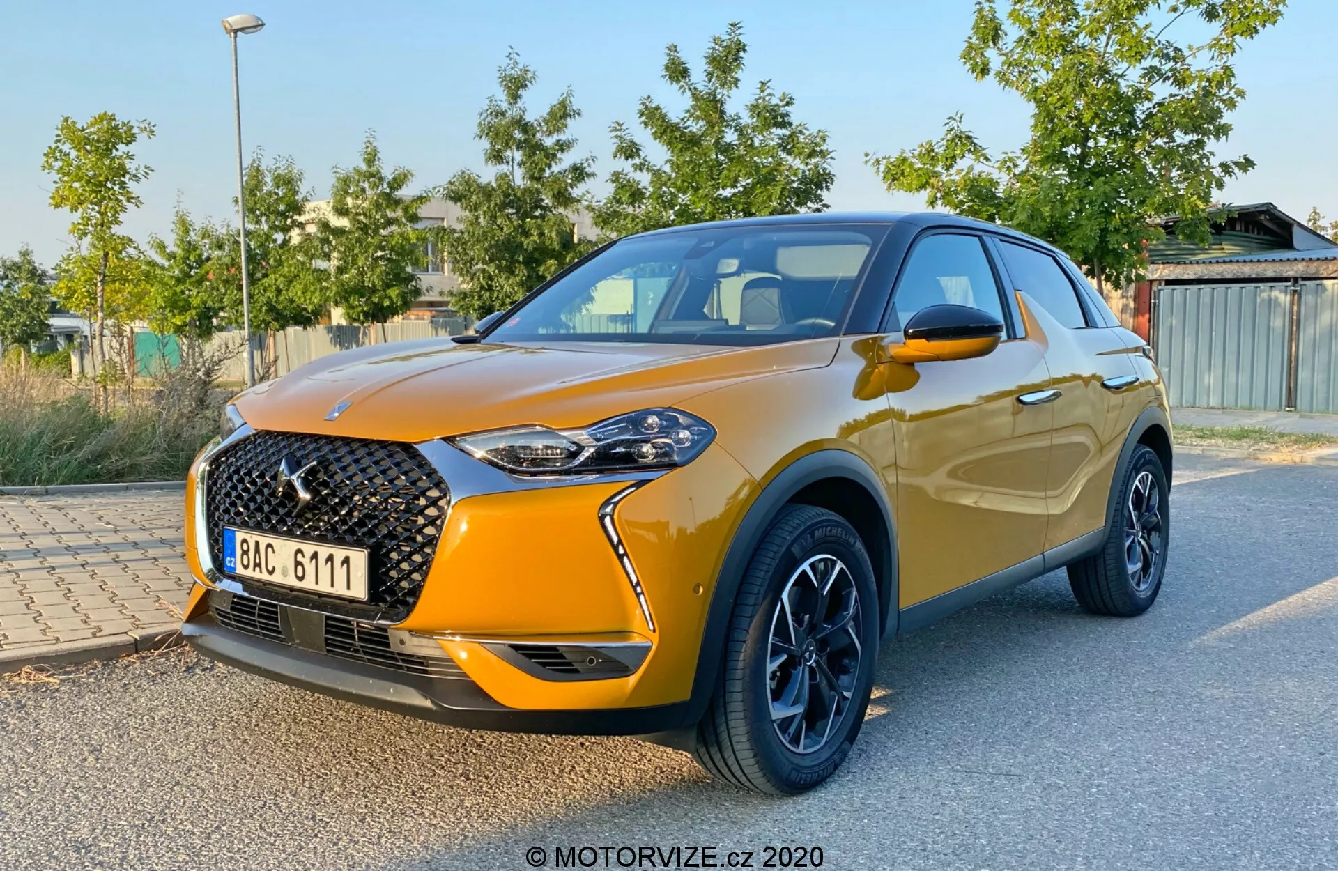 Vista anteriore a tre quarti della DS Automobiles DS 3 Crossback 2019, che mostra la distintiva grande griglia anteriore con motivo a rete e badge prominente, eleganti fari LED, verniciatura metallizzata arancione, cerchi in lega neri a contrasto, specchietti laterali integrati e linea ascendente dei finestrini che termina con uno spoiler aerodinamico. Il veicolo è parcheggiato con fogliame verde e strutture sullo sfondo sfocato, enfatizzando il design e le caratteristiche sportive dell'auto.