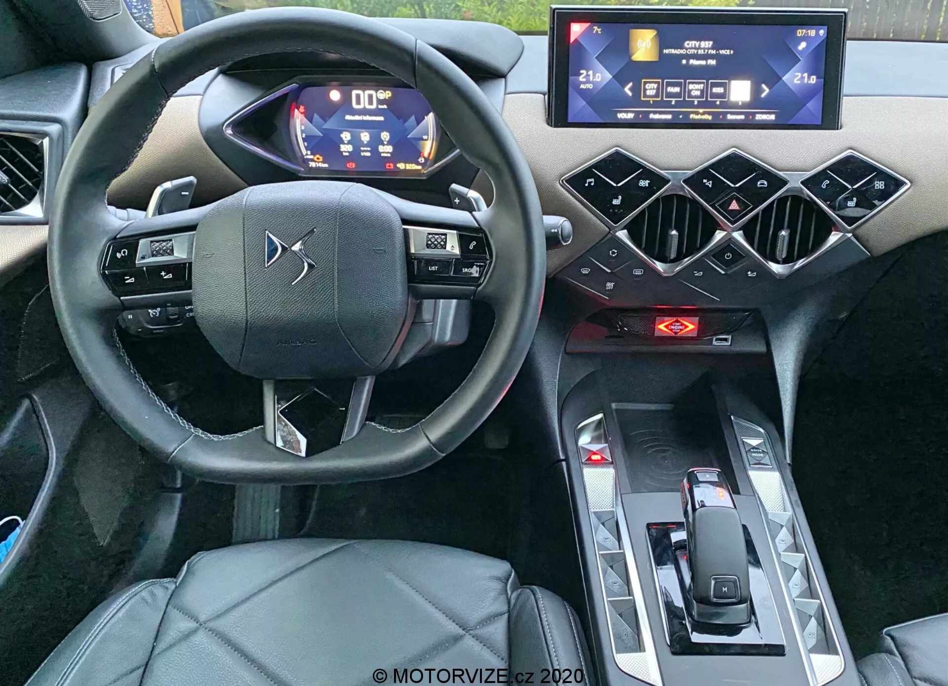 Vista interna della DS Automobiles DS-3 Crossback 2019, focalizzata sul posto di guida da una prospettiva a livello occhi. L'immagine, scattata dall'area del passeggero posteriore, mette in evidenza il volante a tre razze con controlli touch capacitivi, un quadro strumenti digitale con indicatori illuminati in blu e bianco, un sistema di infotainment touchscreen orizzontale, elementi di design esagonali geometrici sulla console centrale, indicatori rossi del controllo del clima, un selettore del cambio minimalista e il sedile del conducente rivestito in pelle o similpelle. La foto enfatizza le caratteristiche moderne, tecnologiche e il design di alta gamma dell'auto.