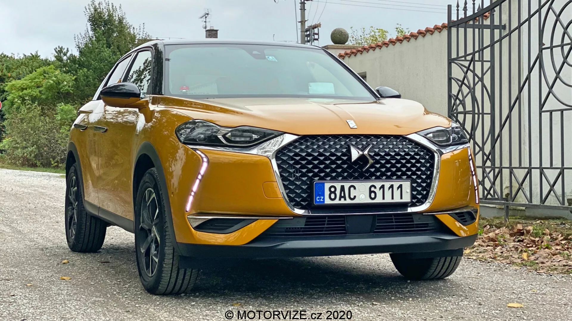 Vista anteriore a tre quarti di un SUV DS Automobiles DS 3 Crossback 2019, che mostra la sua altezza e la robustezza su sfondo suburbano. Sono visibili la vernice metallica dorata, la griglia prominente con motivo a diamante e stemma, i fari LED eleganti, i fendinebbia, il paraurti anteriore di grandi dimensioni con ampia presa d'aria, i cerchi in lega neri a più razze e gli specchietti laterali con indicatori di direzione integrati.