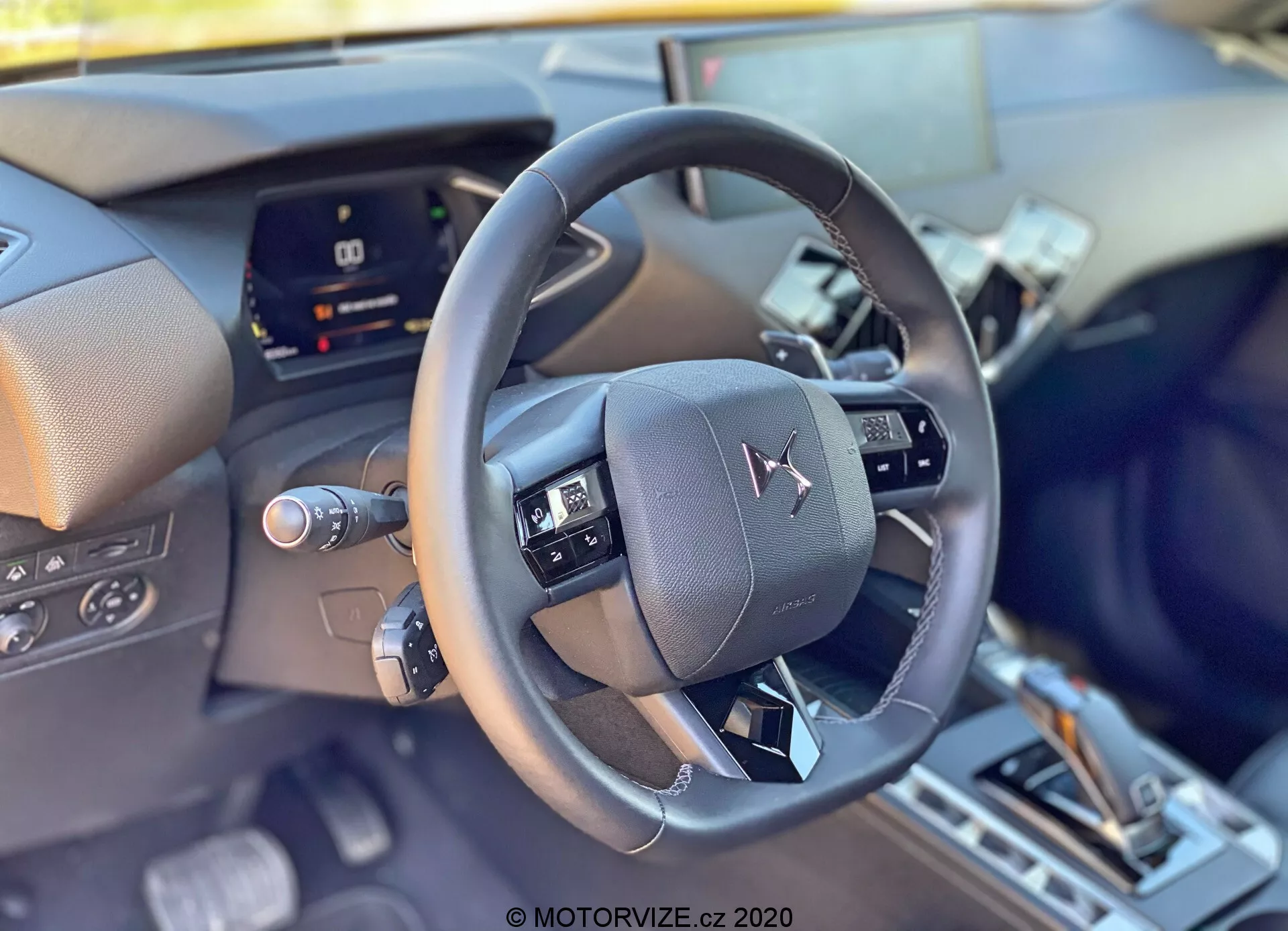 Vista interna ravvicinata della DS Automobiles DS-3 2019 (DS-3 Crossback) dalla prospettiva del conducente, focalizzata sul volante moderno in pelle al centro dell'immagine con comandi multifunzione. Dietro il volante c'è un quadro strumenti digitale, parzialmente oscurato, che indica tecnologia avanzata del veicolo. La parte inferiore suggerisce una console centrale con una leva del cambio e possibili selettori delle modalità di guida, riflettendo caratteristiche interne e di comfort sofisticate.