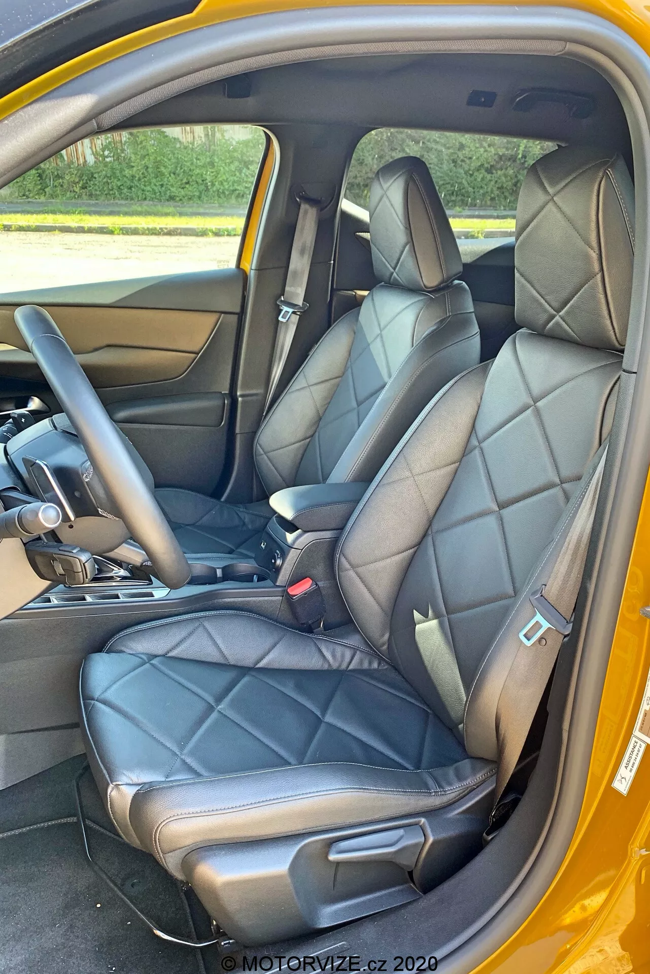 Vista interna di una DS Automobiles DS-3 Crossback 2019 concentrandosi sui sedili anteriori dalla prospettiva della porta laterale del passeggero. L'immagine mette in risalto i sedili in pelle nera trapuntata con poggiatesta integrati e supporti laterali, suggerendo un design lussuoso o sportivo. Parte della console centrale con una leva del cambio che suggerisce una trasmissione automatica è visibile, così come la parte inferiore del cruscotto. Il pannello interno della porta corrisponde alla pelle dei sedili, con una maniglia delle porte in metallo e pulsanti di controllo. Si vedono anche le cinture di sicurezza nere con un meccanismo di regolazione dell'altezza, un pavimento in moquette e uno schema di colore esterno contrastante, possibilmente giallo.