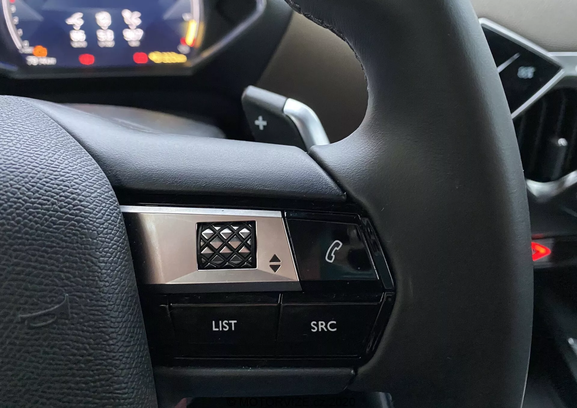 Vista ravvicinata dalla prospettiva del conducente del lato sinistro del volante in un DS Automobiles DS 3 Crossback 2019. L'immagine mette a fuoco i pulsanti di controllo, compreso un pulsante con icona del telefono, un pulsante 'LIST' e un pulsante 'SRC', evidenziati contro uno sfondo sfocato del cruscotto. Il design ad alto contrasto combina finiture nere e metalliche con un motivo a diamante sul cromo, migliorandone l'aspetto premium.
