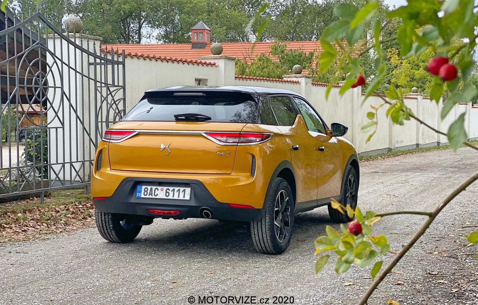 Vista angolare posteriore di una DS Automobiles DS 3 Crossback 2019, catturata da una prospettiva bassa su un vialetto residenziale. L'auto presenta lampeggianti posteriori orizzontali prominenti, logo del produttore centrato su un portellone scolpito con spoiler integrato e terza luce di stop, targa centrale e finitura metallica lucida di colore arancione o dorato. Il robusto paraurti posteriore ha sensori o riflettori integrati, con tergicristallo posteriore visibile, antenna a pinna di squalo e ruote in lega a più raggi. La vegetazione leggermente sfocata in primo piano contrasta con la messa a fuoco più nitida sull'auto e sul cancello di ingresso sullo sfondo, suggerendo una luce diurna morbida e diffusa.