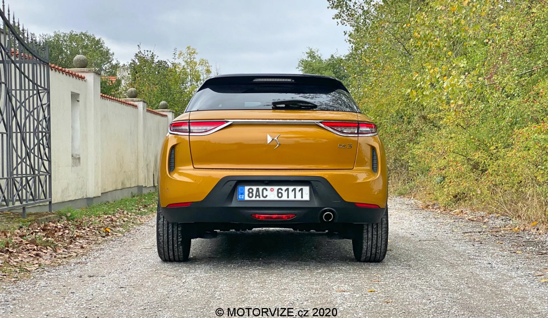 Vista posteriore di una moderna DS-Automobiles DS-3 Crossback 2019 che mostra il design e le caratteristiche tecnologiche, incluso lo spoiler montato sul tetto, il parabrezza posteriore con elemento riscaldante, la luce di stop montata in alto, il cofano del bagagliaio con logo del produttore, la striscia cromata prominente, i fanali posteriori LED eleganti, il paraurti posteriore robusto con sensori di parcheggio, il sistema di scarico doppio, l'elemento simile al diffusore nero e un colore arancione o rame accattivante su una superficie ghiaiosa con vegetazione sullo sfondo.