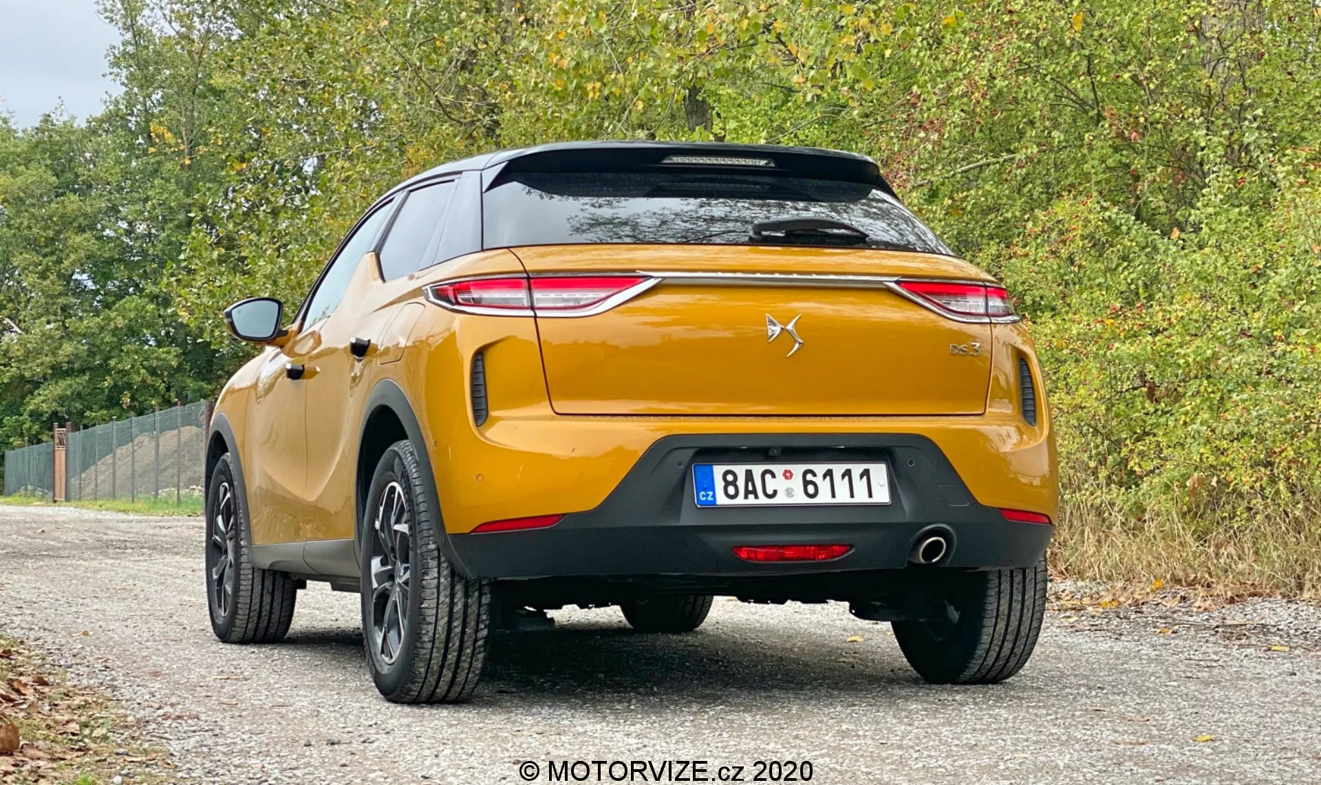 L'immagine mostra una vista posteriore di una moderna DS-3 Crossback 2019 dorata, parcheggiata su una strada sterrata con vegetazione su entrambi i lati. L'auto, catturata da un angolo basso, presenta i fari posteriori affilati diretti verso il centro del bagagliaio, un emblema stilizzato a forma di chevron e leone sul portellone, un robusto paraurti posteriore con sensori di parcheggio, doppi scarichi, rifiniture in plastica nera e cerchi in lega a cinque razze. La targa è in stile europeo, con riflettori posteriori standard e un fendinebbia posteriore. L'illuminazione morbida e diffusa suggerisce una giornata nuvolosa, aggiungendo all'atmosfera casual e versatile, mentre la filigrana ‘MOTORVIZE.cz 2020’ indica la fonte dell'immagine.