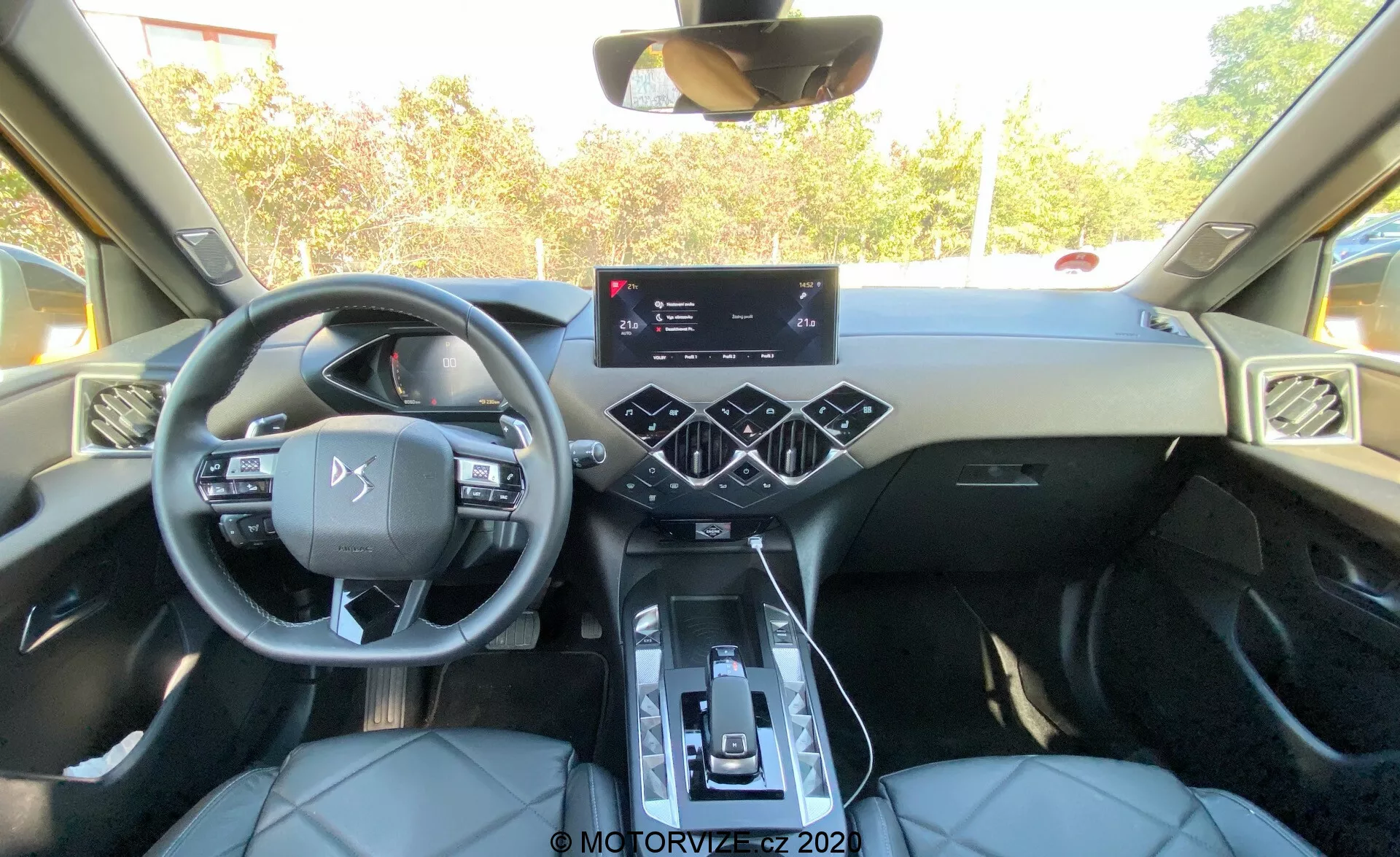 Interno di una moderna DS Automobiles DS-3 2019 (DS-3 Crossback) visto dalla prospettiva del sedile posteriore, che mostra l'area della cabina anteriore con cruscotto, volante, console centrale con cambio, portabicchieri, pannello di controllo del clima touch, touchscreen di infotainment prominente, volante multifunzione, bocchette d'aria angolari, sedili anteriori in pelle con cuciture a contrasto, rifiniture della porta con pulsanti di controllo e tetto panoramico.