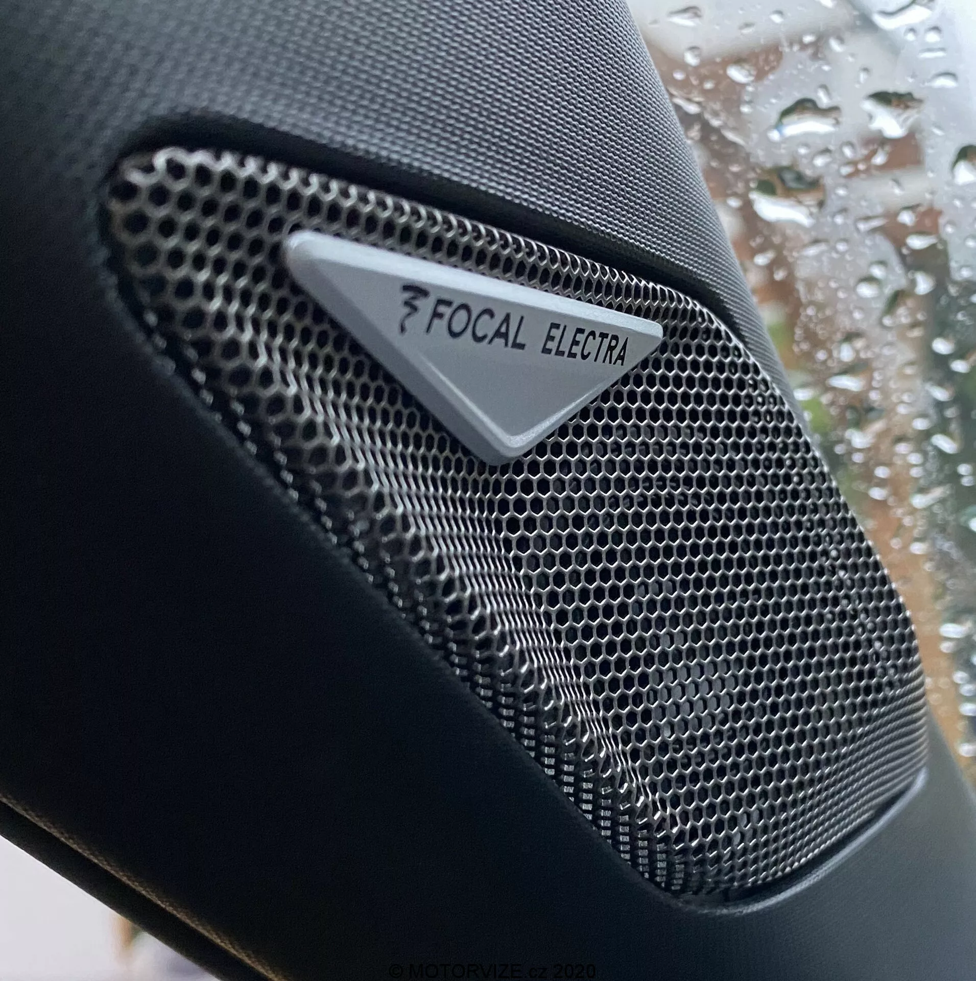 Primo piano dell'altoparlante con il marchio Focal Electra nella DS Automobiles DS-3 Crossback 2019, che mostra il componente audio ad alta fedeltà con una griglia a nido d'ape in metallo di alta qualità e un chiaro logo Focal e marchio Electra. La foto si concentra sul design intricato dell'altoparlante, con una bassa profondità di campo che mette in risalto la texture dell'altoparlante contro uno sfondo sfocato di un finestrino dell'auto con gocce di pioggia.