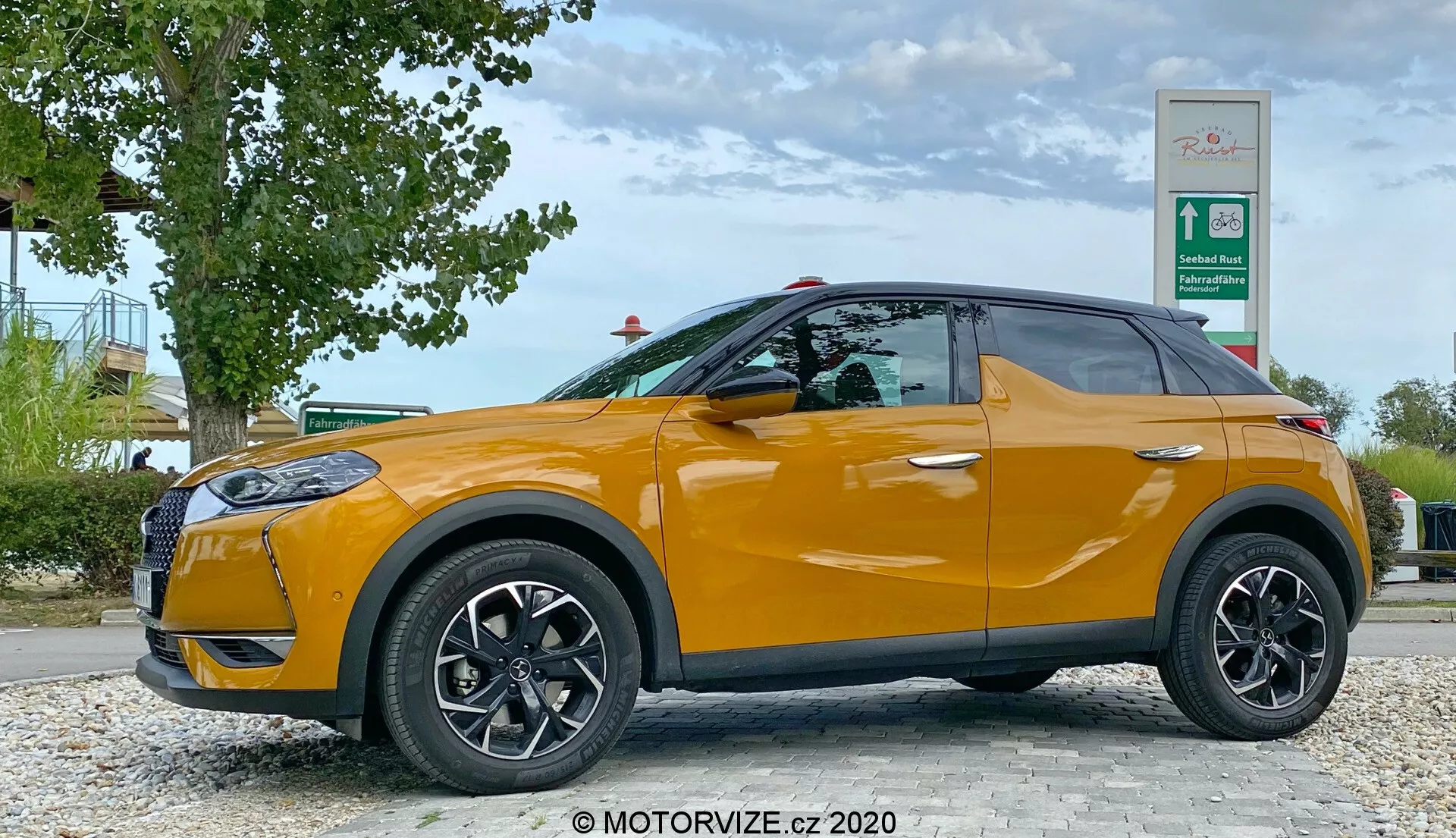 Profilo laterale di una DS Automobiles DS 3 Crossback 2019 arancione brillante, parcheggiata vicino a una zona stradale con vegetazione e un'insegna di stazione di servizio. Il lato del conducente, con ruote nere a più razze e specchietti laterali dello stesso colore della carrozzeria, è visibile da un angolo basso all'altezza delle ruote dell'auto, mostrando il suo design elegante di SUV compatto con una linea di cintura ascendente e finestrini oscurati.