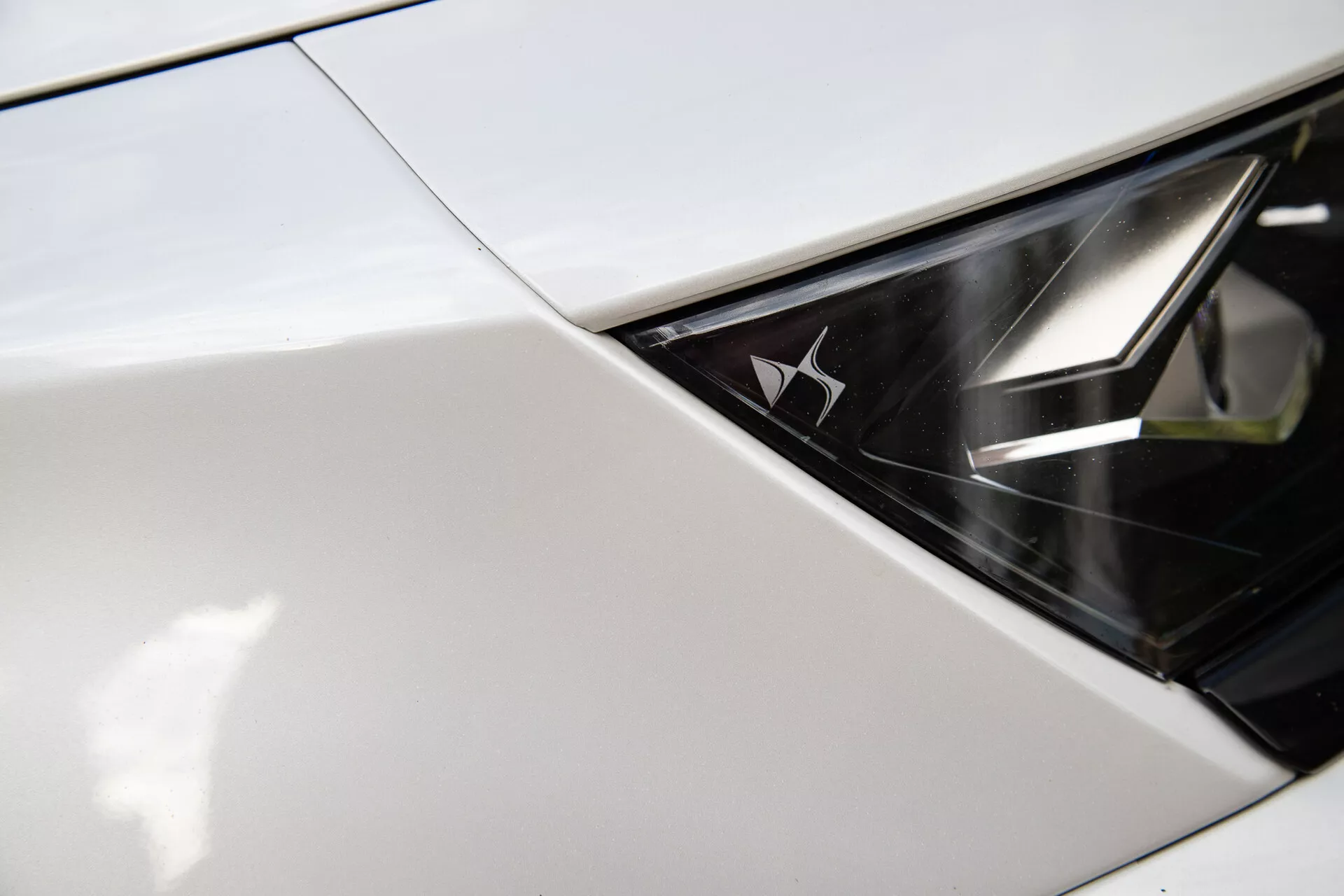 Immagine dettagliata del faro della DS Automobiles DS-7 2022 che mostra lo scontro tra il parafango o il cappuccio con l'insieme del faro, esibendo un design lucido e monocromatico e un emblema del marchio dentro il faro, suggerendo lusso moderno e tecnologia avanzata di illuminazione LED.