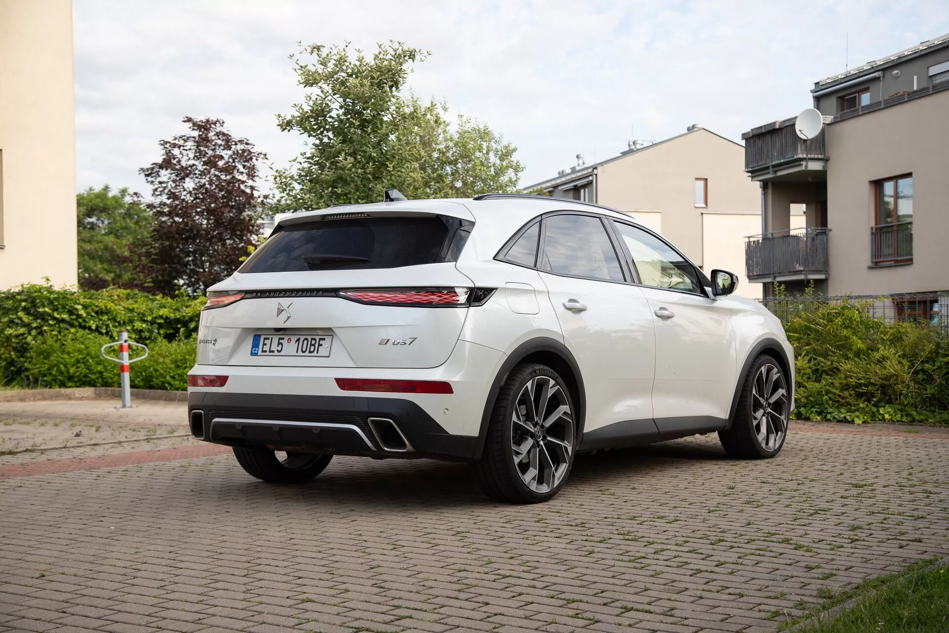Vista posteriore tre quarti di una DS Automobiles DS 7 2022 in bianco, mostrando il profilo laterale e posteriore con distintive luci posteriori orizzontali sottili, badging, spoiler alto montato, antenna a pinna di squalo, sensori di parcheggio, sistema di scarico doppio e grandi ruote in lega a più razze su una strada residenziale.