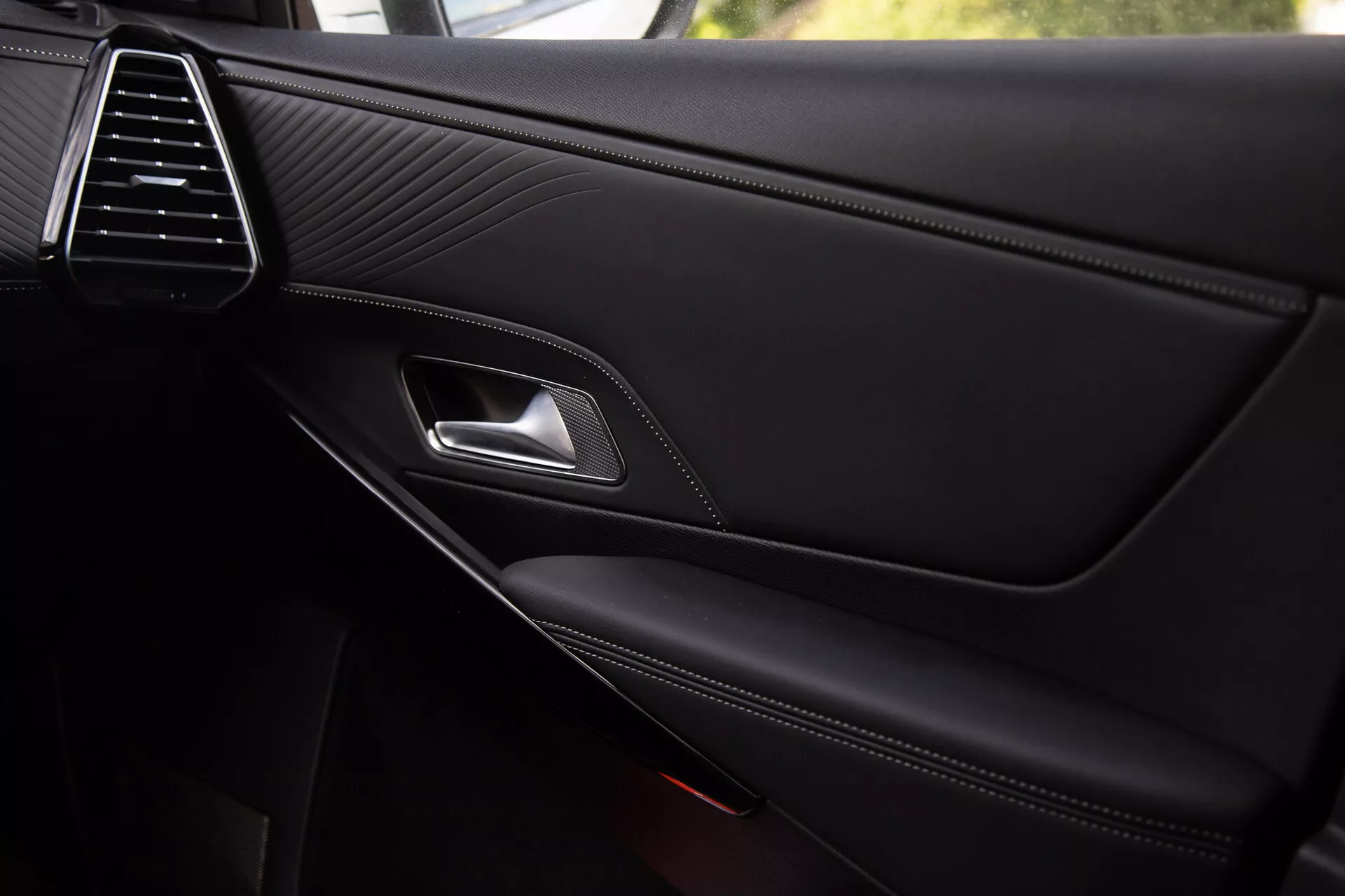 Vista ravvicinata del pannello della porta interna della DS Automobiles DS-7 2022 che mette in mostra la sofisticata bocchetta d'aria, la texture in pelle cucita, la maniglia della porta in cromo, la leva di sgancio ergonomica, il bracciolo con cuciture a contrasto e la tasca di stoccaggio integrata con griglia dell'altoparlante parzialmente visibile, riflettendo una qualità premium e dettagli di design.