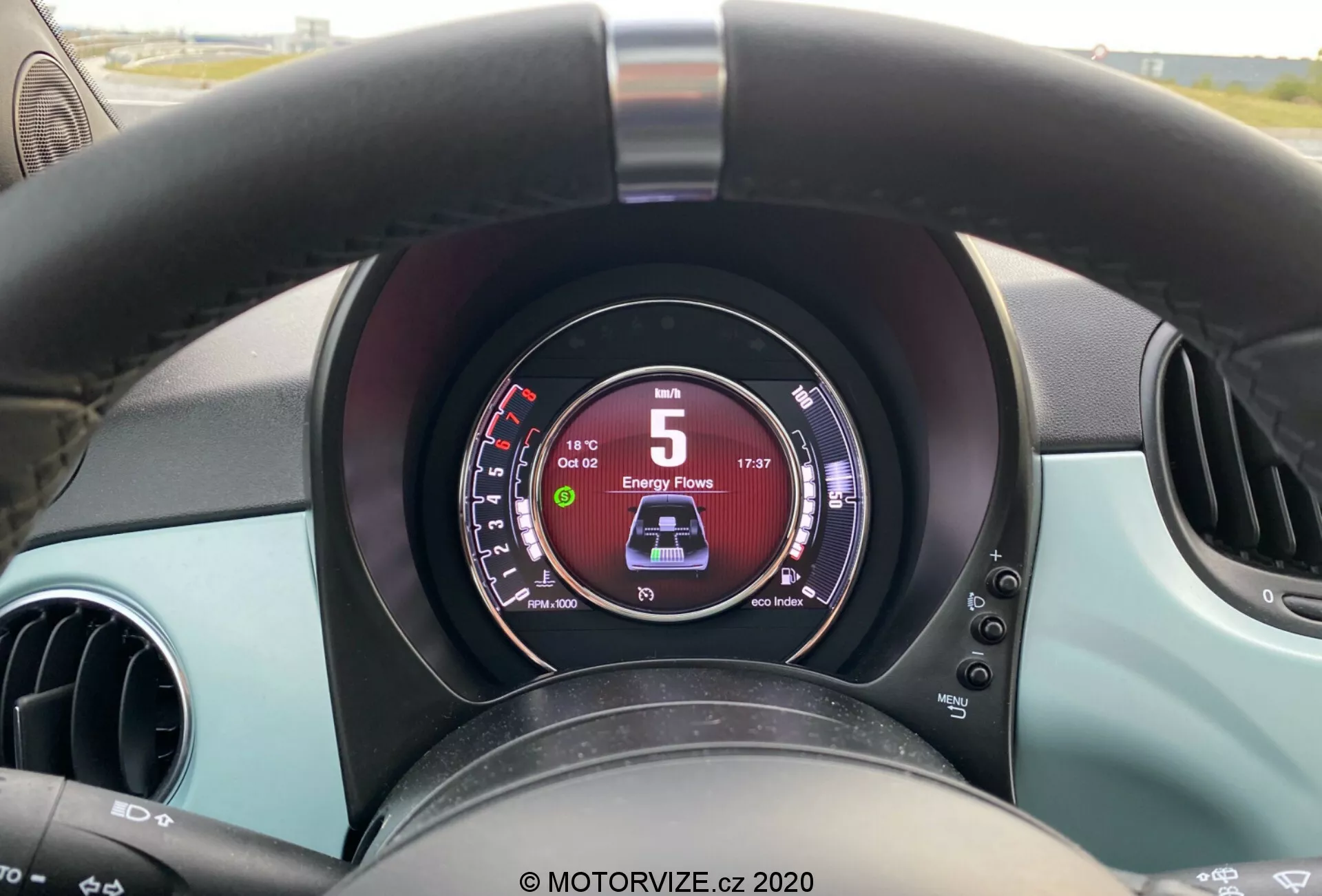 Vista ravvicinata dalla prospettiva del conducente dell'interno di una Fiat 500 2015, con in evidenza un volante nero leggermente usurato, un cruscotto digitale che mostra 5 km/h e 'D' per la guida, due prese d'aria circolari e accenti turchesi sul cruscotto e sulle porte.