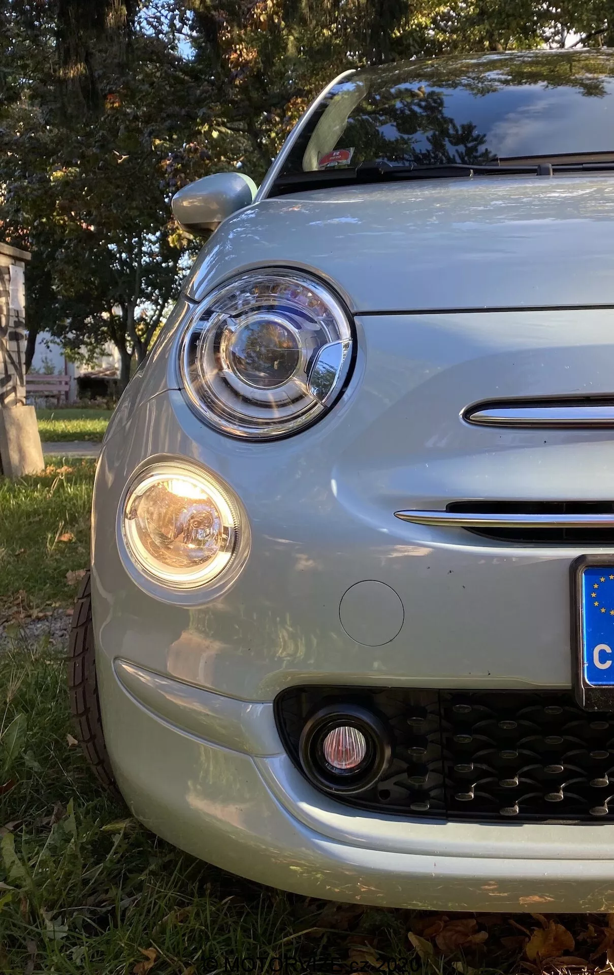 Vista ravvicinata del quarto anteriore sinistro di una Fiat 500 2015 ripresa da un'angolazione bassa, che mette in risalto il cofano, il paraurti anteriore, il parafango e il faro principale circolare con proiettore circondato da un anello DRL. Il cluster include una freccia di svolta ambrata e un fendinebbia sotto. Accenti cromati adornano il bordo del faro, l'alloggiamento del fendinebbia e le due barre orizzontali della griglia. La griglia presenta un pattern a nido d'ape nero. Il frontale arrotondato e la targa in stile europeo suggeriscono il design retro-moderno dell'auto, parcheggiata sull'erba con foglie autunnali.