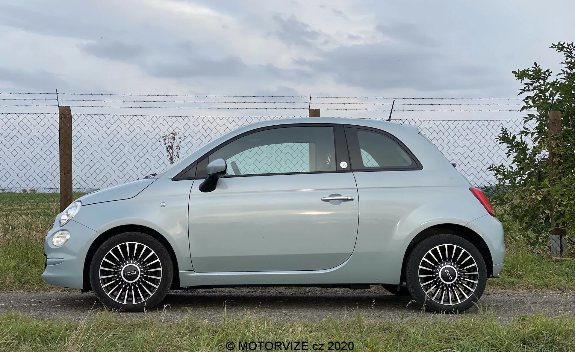 Moderna autovettura hatchback subcompatta in un’angolazione di profilo laterale, che mostra l'intera lunghezza del veicolo. Parcheggiata su una superficie piana con una recinzione perimetrale e un orizzonte con nuvole sullo sfondo. Caratteristiche: porte laterali, finestrini, specchietti, configurazione a due porte, piccolo finestrino posteriore, silhouette del tetto arrotondata, colore della carrozzeria simile a un pastello con tetto in contrasto, cerchi in lega a raggi multipli, pneumatici in buone condizioni, linee di design dettagliate e illuminazione naturale diurna.