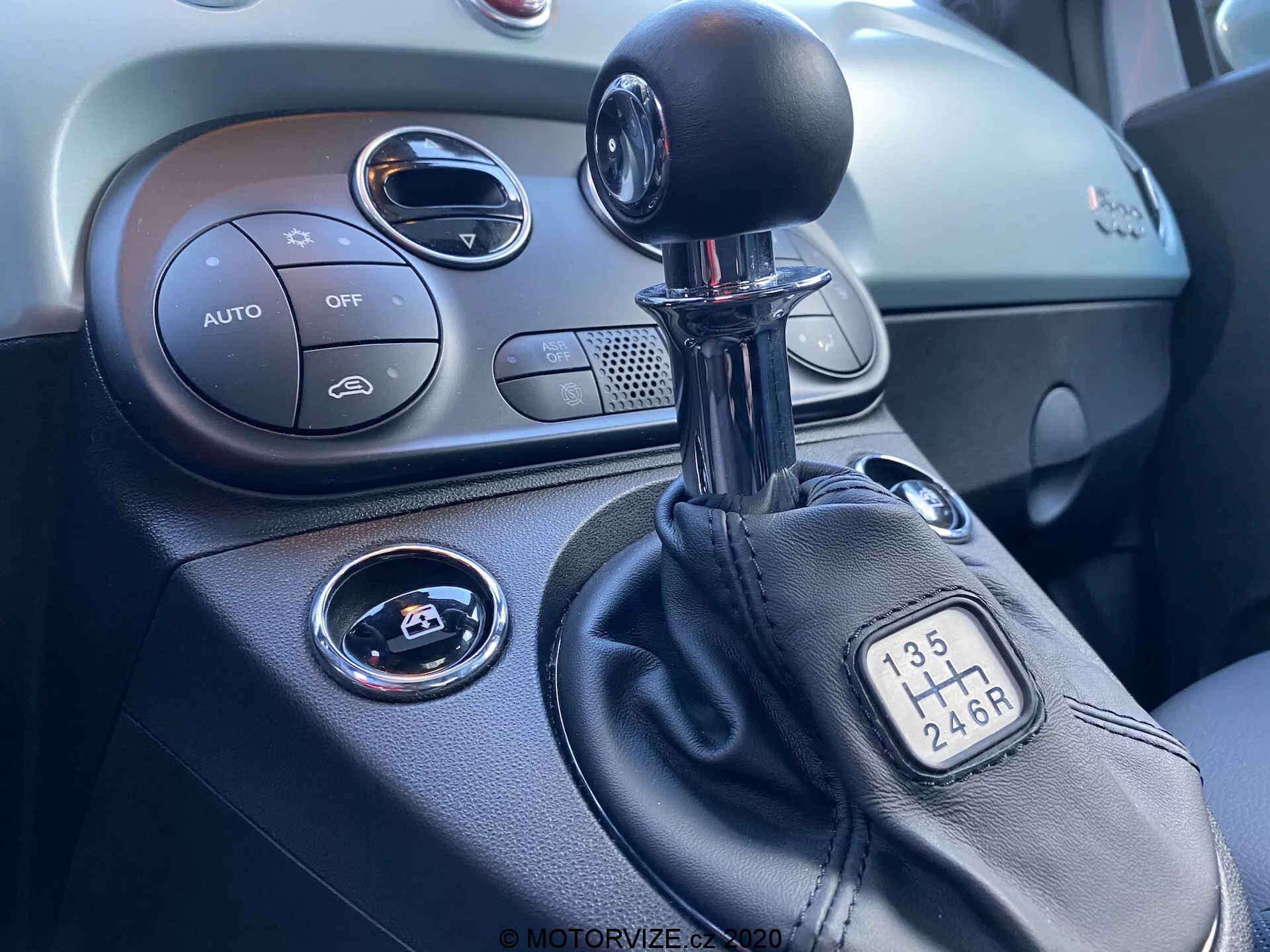 Vista ravvicinata della console centrale all'interno di un Fiat 500 del 2015, focalizzata sul lato destro. L'immagine mostra una leva del cambio manuale con configurazione a 6 marce, parte della console centrale con comandi del climatizzatore etichettati 'AUTO', pulsante 'OFF', pulsante start/stop per l'accensione, presa d'aria circolare e il cruscotto testurizzato.