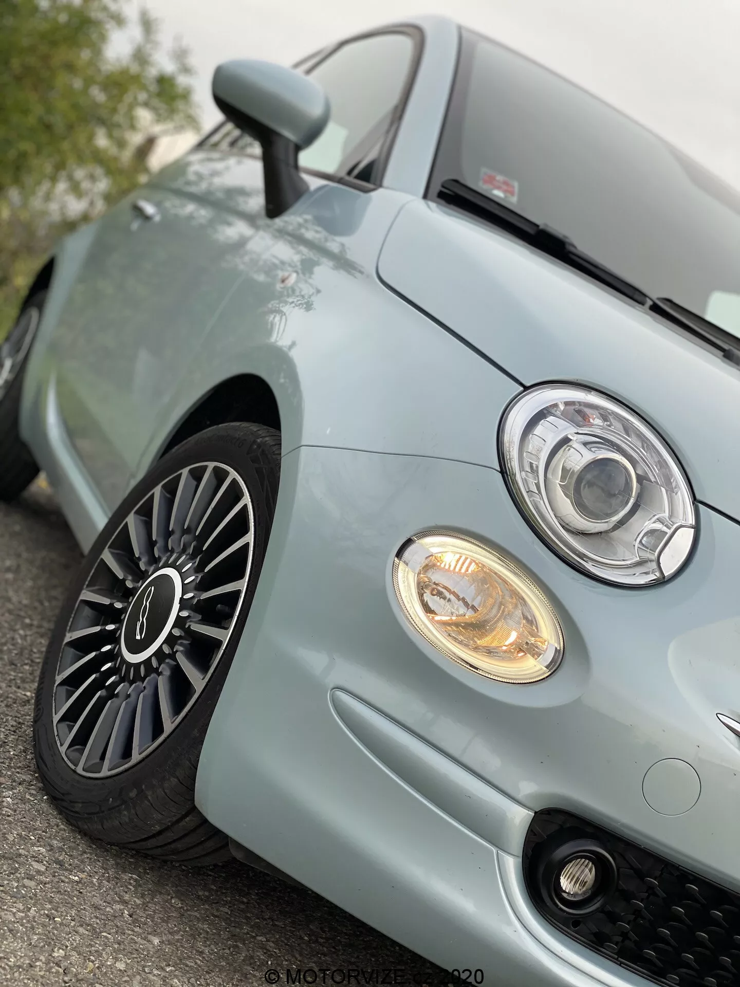 Vista ravvicinata e angolata dal basso del quarto anteriore sinistro di una Fiat 500 2015 blu chiaro, che mostra il faro moderno in stile proiettore con anello LED o halo, il fendinebbia illuminato, la griglia sportiva a nido d'ape, la sezione anteriore del cofano e la ruota in lega multirazze con buona profondità del battistrada, suggerendo un veicolo ben mantenuto e un avanzato design automobilistico.