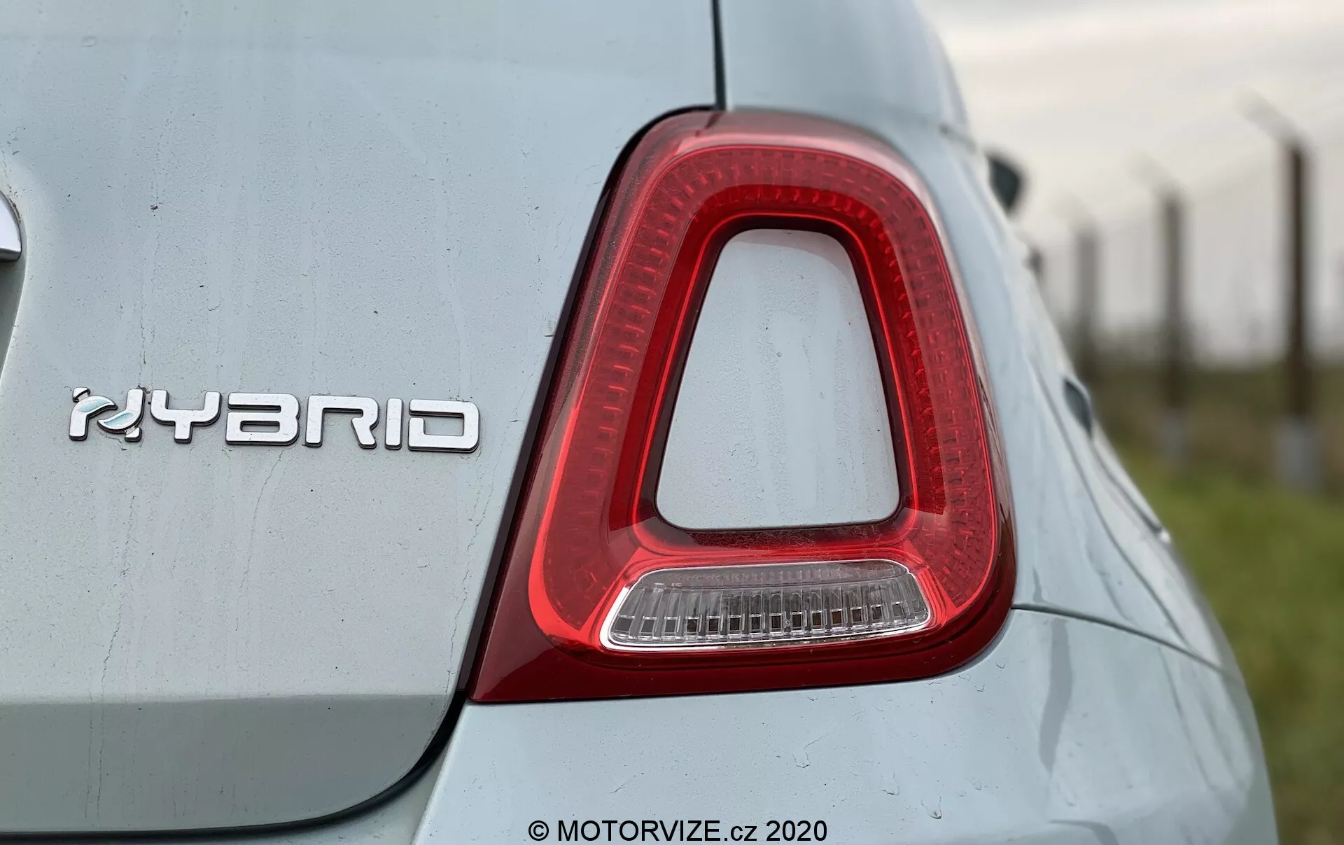 Dettaglio ravvicinato del fanale posteriore destro e del badge 'HYBRID' su una Fiat 500 2015. Il fanale ha una forma a L rovesciata, prevalentemente rosso con una sezione trasparente per la luce di retromarcia ed è a filo con la carrozzeria. Il badge metallico 'HYBRID', indicativo del sistema ibrido, è posizionato in modo prominente. Il colore della carrozzeria, chiaro, presenta una qualità riflettente con alcune macchie di polvere o acqua, suggerendo un ambiente esterno. L'immagine è presa da un'angolazione leggermente bassa, evidenziando il design del fanale e la tecnologia ibrida.