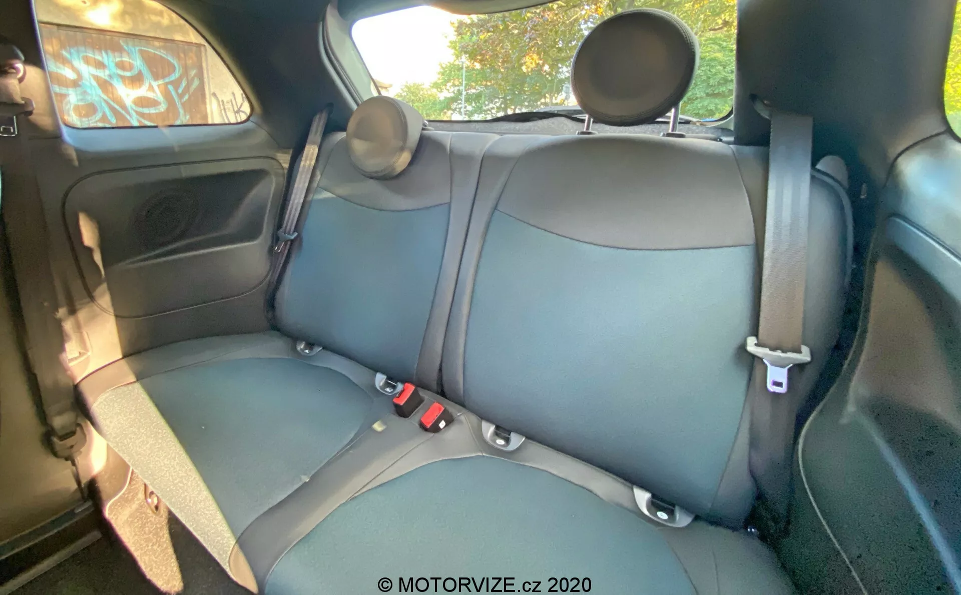 Interni di una Fiat 500 del 2015 che mostrano i sedili posteriori con rivestimento in pelle o similpelle di colore scuro, cuciture visibili, cinture di sicurezza a tre punti, poggiatesta regolabili, pannello della porta con controllo elettrico del finestrino, vano portaoggetti, griglia dell'altoparlante, vista parziale del soffitto e maniglia superiore. L'angolo della telecamera è leggermente elevato da davanti a dietro, fornendo una vista dettagliata del sedile posteriore, della porta posteriore, di parte del soffitto e dei finestrini.