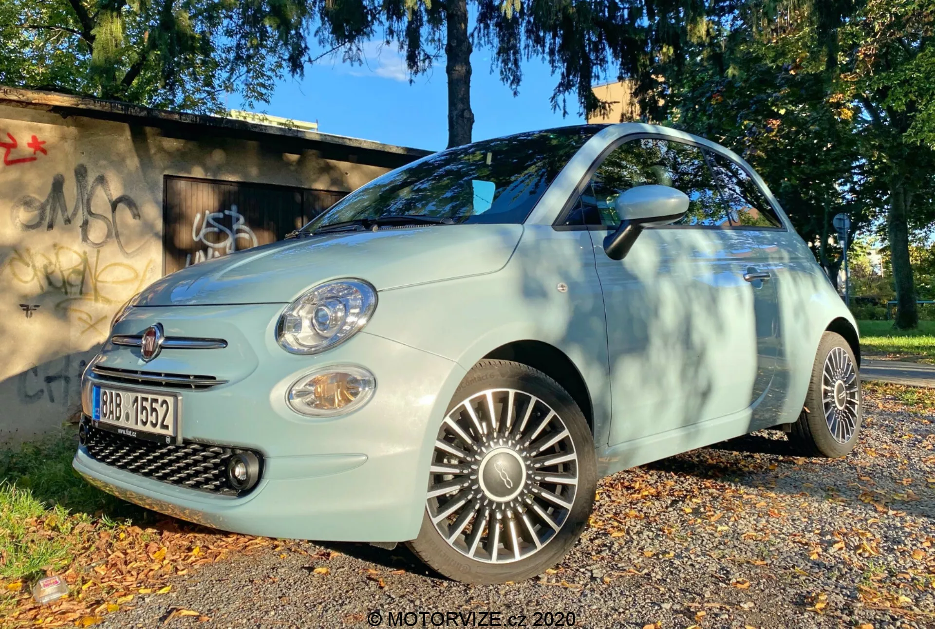 Scatto a bassa angolazione di una Fiat 500 2015 dal quarto frontale, che mostra il lato del conducente. L'immagine mette in evidenza la griglia, il paraurti anteriore, i fanali, i fendinebbia e le ruote in lega dettagliate. L'auto è parcheggiata su una superficie coperta da foglie autunnali, con un'area erbosa e una struttura graffitata sullo sfondo, illuminata dalla luce naturale del giorno, suggerendo un'illuminazione del mattino o del tardo pomeriggio.