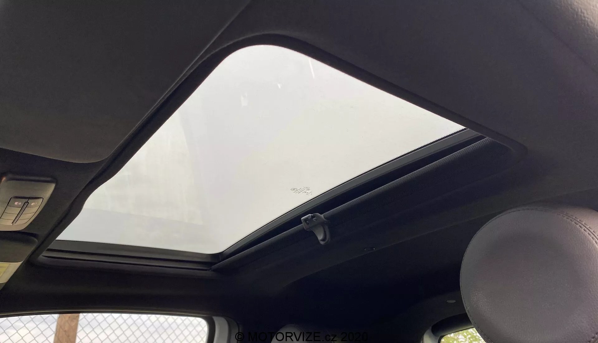 Vista interna di una Fiat 500 2015 con focus sulla sezione superiore verso il tetto dalla prospettiva di un passeggero o conducente. Il tetto apribile chiuso, prominente, permette alla luce naturale di entrare nell'abitacolo. Il rivestimento del tetto scuro contrasta con l'esterno. Maniglia visibile sul lato sinistro, luce standard di cupola sopra e i bordi di due visiere parasole in alto. Un gancio appendiabiti è vicino al bordo del vetro del tetto apribile, che ha un'etichetta di sicurezza nella parte inferiore. L'immagine è scattata da un angolo basso, inquadrando da vicino le caratteristiche del tetto interno.