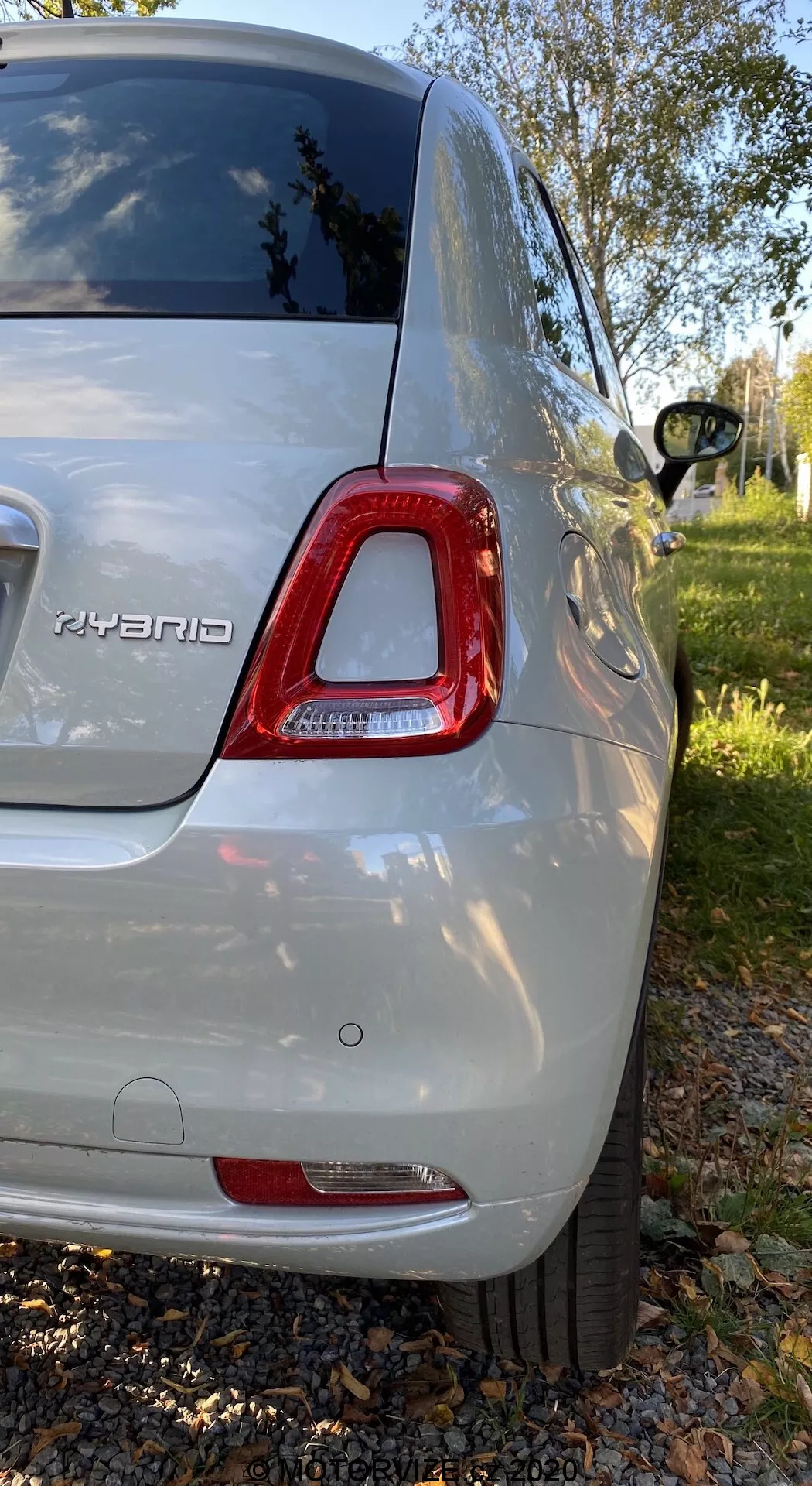 Vista posteriore destra di una Fiat 500 ibrida del 2015 in colore argento metallizzato, che mostra il fanale posteriore a LED, il badge ibrido, il paraurti posteriore, la ruota parzialmente visibile, il finestrino posteriore oscurato e la carrozzeria brillante; fotografato in un'ambientazione suburbana con vegetazione e alberi.