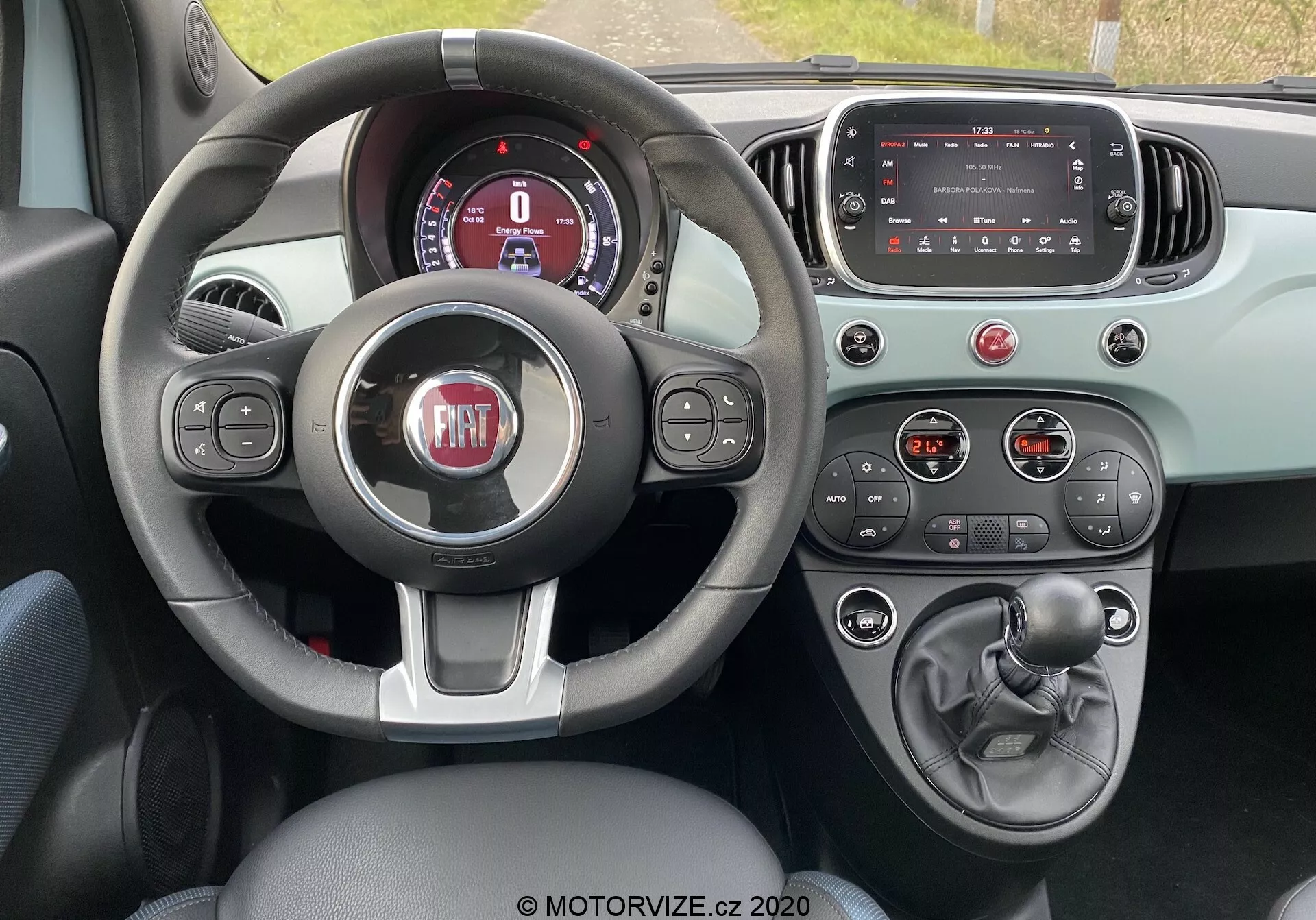 Vista interna della Fiat 500 2015 dalla prospettiva del conducente che mostra il volante a tre razze con pulsanti di controllo, cruscotto digitale parzialmente visibile, schermo touchscreen del sistema di infotainment spento, pulsanti e manopole per il controllo del clima, leva del cambio manuale, freno a mano e schema cromatico bicolore che enfatizzano funzionalità ed elementi di design moderno.