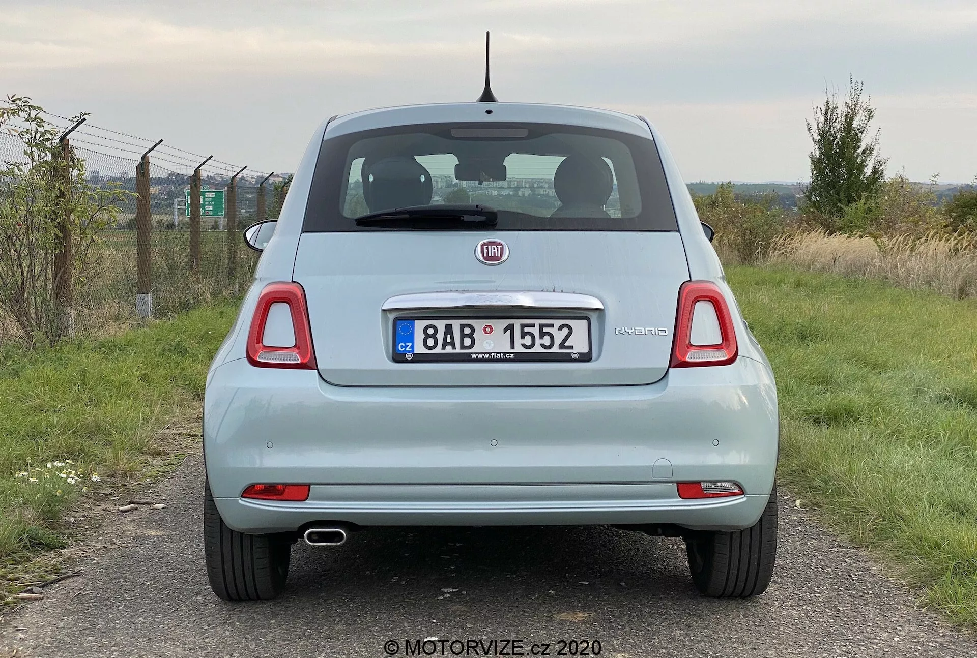 Vista posteriore di una Fiat 500 del 2015 di colore chiaro, scattata ad altezza occhi e che mostra l'intera larghezza del veicolo. L'immagine mette in evidenza il portellone con il logo del marchio e il badge del modello, il paraurti posteriore, le luci posteriori, il tergilunotto posteriore e i doppi tubi di scarico. Sono visibili anche i riflettori posizionati vicino agli angoli del paraurti e l'antenna montata sul tetto. La targa e l'ambiente circostante sono visibili parzialmente, ma il punto focale rimane il veicolo.