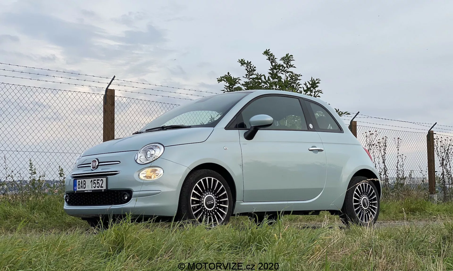 Foto a bassa angolazione di una Fiat 500 del 2015, evidenziando la sua posizione robusta. Vista laterale e parziale anteriore, con fari rotondi, dettagli cromati e stemma visibile. Veicolo a due porte colore pastello parcheggiato su un'area erbosa con sfondo industriale o semi-urbano. Ruote dal design multirazze, specchietti laterali color carrozzeria e antenna posteriore visibile. Griglia anteriore modesta con badge Fiat in cromo.