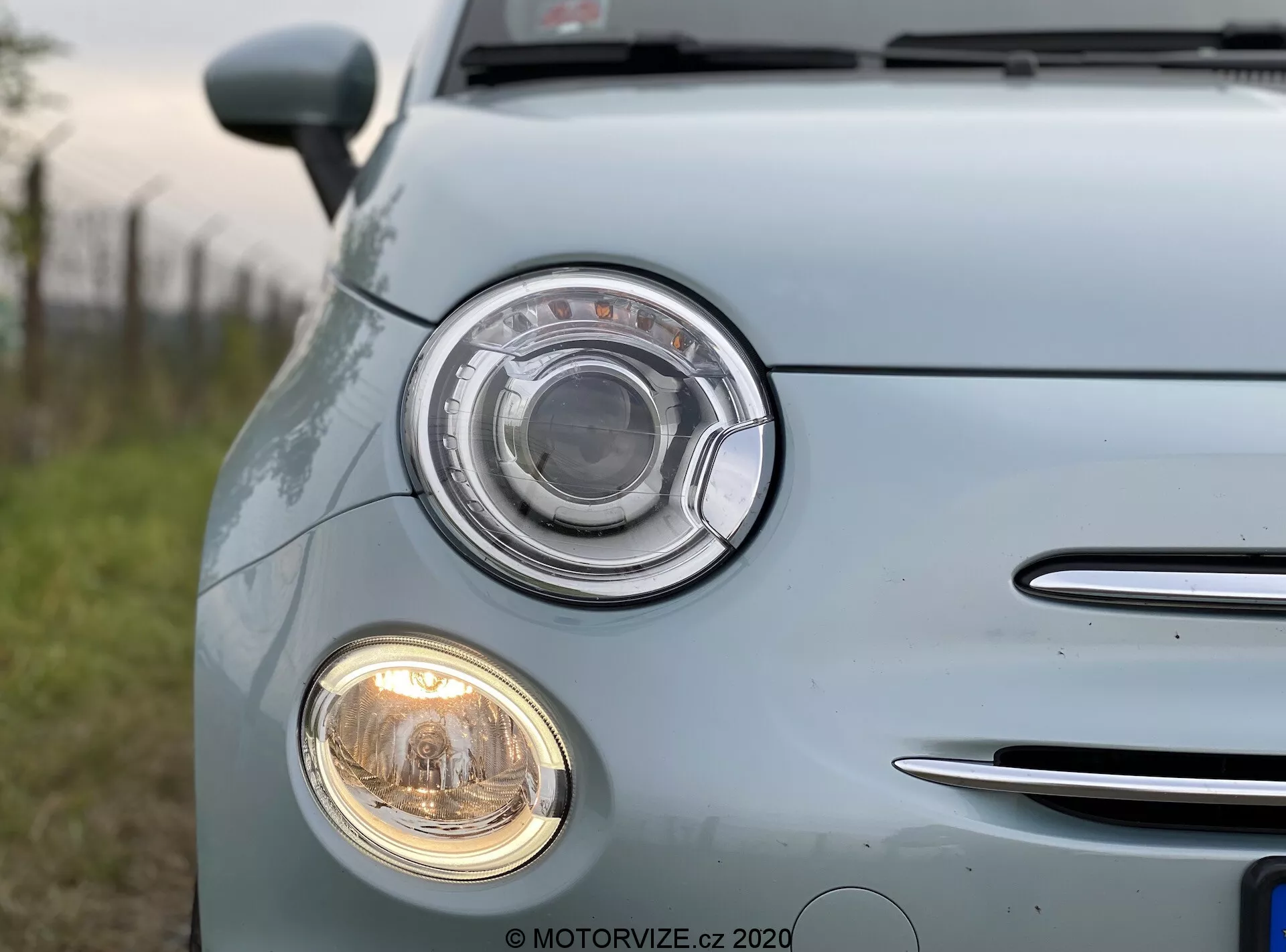 Immagine ravvicinata di una Fiat 500 2015 blu chiaro ripresa dal quarto anteriore sinistro. Il focus è sull'assemblaggio dei fari distintivo con un design circolare e lente trasparente, così come sul faro fendinebbia circolare più piccolo sotto di esso. Alla sinistra è visibile un segmento della griglia con listelli orizzontali cromati. La vernice blu pallido e lucida dell'auto riflette la luce naturale, mettendo in risalto la sua superficie ben mantenuta. Le insegne e i loghi del veicolo sono stati offuscati.