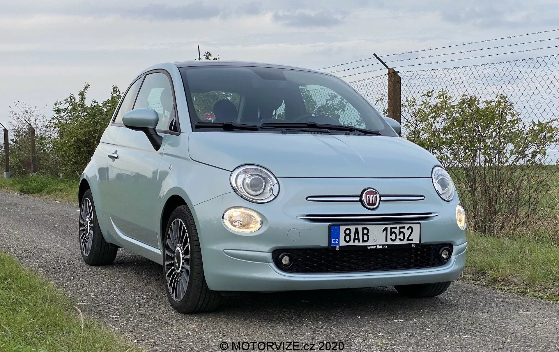 Vista frontale a bassa angolazione di una Fiat 500 del 2015, evidenziando la facciata anteriore, i fari rotondi, i dettagli cromati della griglia, i fari fendinebbia e il distintivo Fiat prominente. Il cofano dell'auto presenta curve caratteristiche e il paraurti anteriore include sensori di parcheggio visibili. Sono visibili anche i cerchi in lega. Il veicolo è parcheggiato sul bordo della strada con un'area erbosa e una recinzione sullo sfondo, indicando un ambiente rurale o semi-rurale.