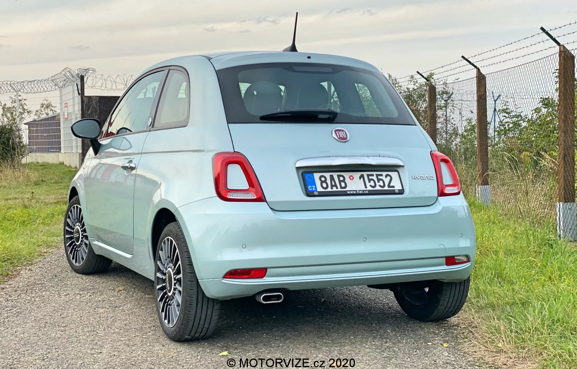 L'immagine mostra una vista posteriore a tre quarti di una Fiat 500 hatchback del 2015 di colore azzurro chiaro. Il fuoco è sul retro e su parte del profilo laterale, presa dal lato posteriore destro del veicolo. Evidenzia la ruota posteriore destra, la luce posteriore, la vista parziale dello specchietto laterale destro e la portiera del passeggero. L'auto è posizionata leggermente a sinistra, con l'angolo della fotocamera leggermente più basso della linea del tetto. Dettagli notevoli includono un singolo scarico, ruote in lega, luci posteriori a lampadina standard, distintivi di marca e modello, un piccolo spoiler posteriore, un'antenna in stile albero e un tappo del carburante visibile. L'ambiente appare rurale con cieli aperti, vegetazione e una recinzione.