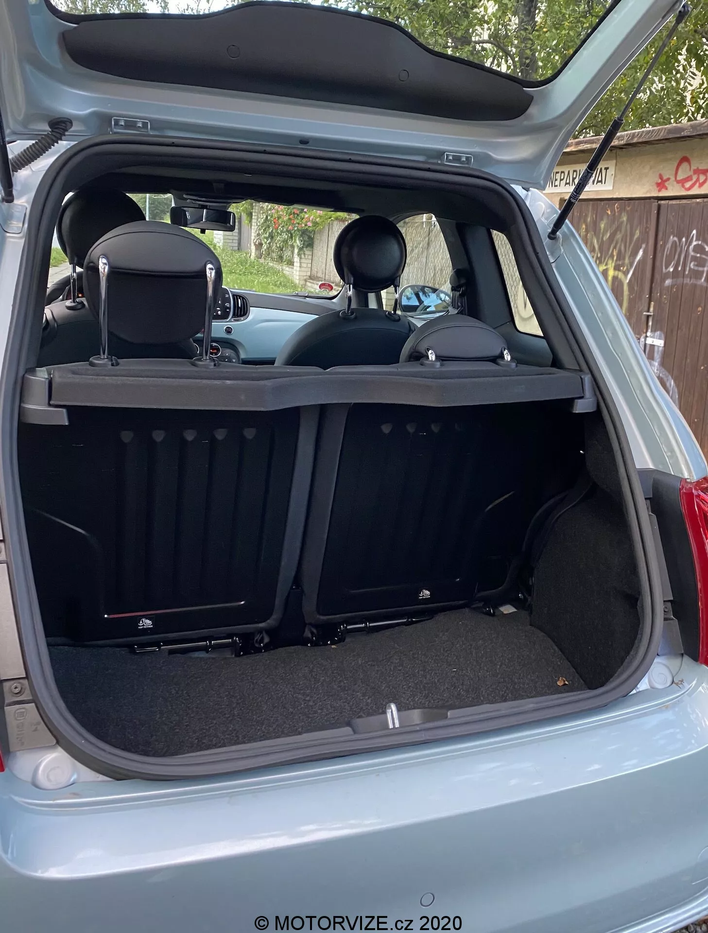 L'immagine mostra l'interno posteriore della Fiat 500 2015 da un'angolazione esterna posteriore con il portellone del bagagliaio completamente aperto. L'area di carico è vista da un angolo leggermente decentrato verso il basso dal lato destro, rivelando la profondità del bagagliaio e varie caratteristiche del retro dell'interno. Il design moderno e pulito include superfici in moquette, una copertura retrattile del vano bagagli a coste, sedili posteriori ribaltabili con poggiatesta e una piastra protettiva. Le caratteristiche tecnologiche includono una luce interna, una presa di corrente da 12V, modanature in plastica, ritagli circolari e pannelli laterali.
