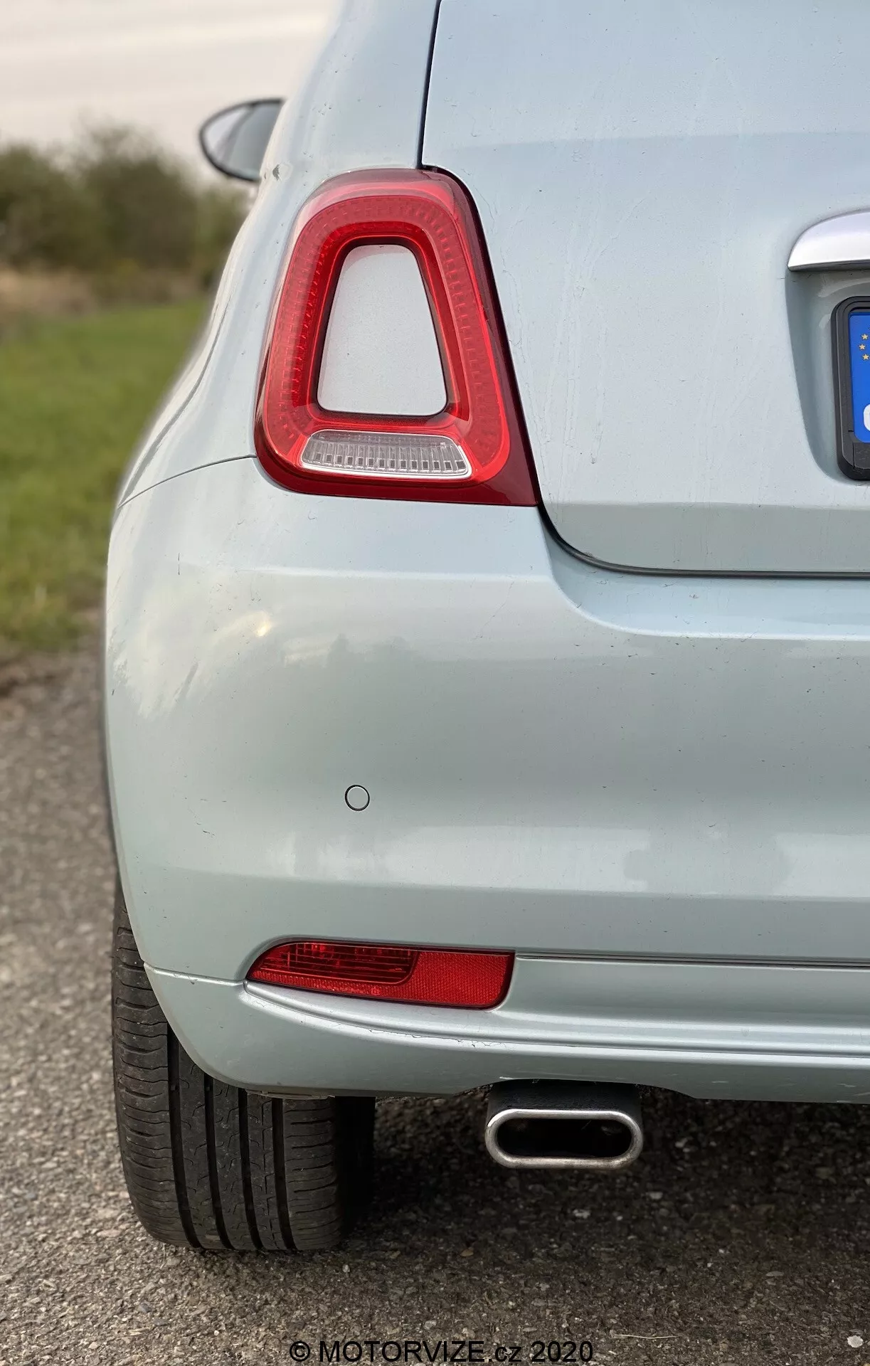 Vista ravvicinata e angolata del lato posteriore sinistro di un'auto di colore chiaro, con particolare attenzione alla caratteristica luce posteriore con lente rossa trasparente e tecnologia LED, riflettore rosso, sensore di parcheggio a filo e tubo di scarico con finitura cromata. La vernice metallizzata chiara e la bassa profondità di campo enfatizzano le buone condizioni di pulizia e manutenzione del veicolo.