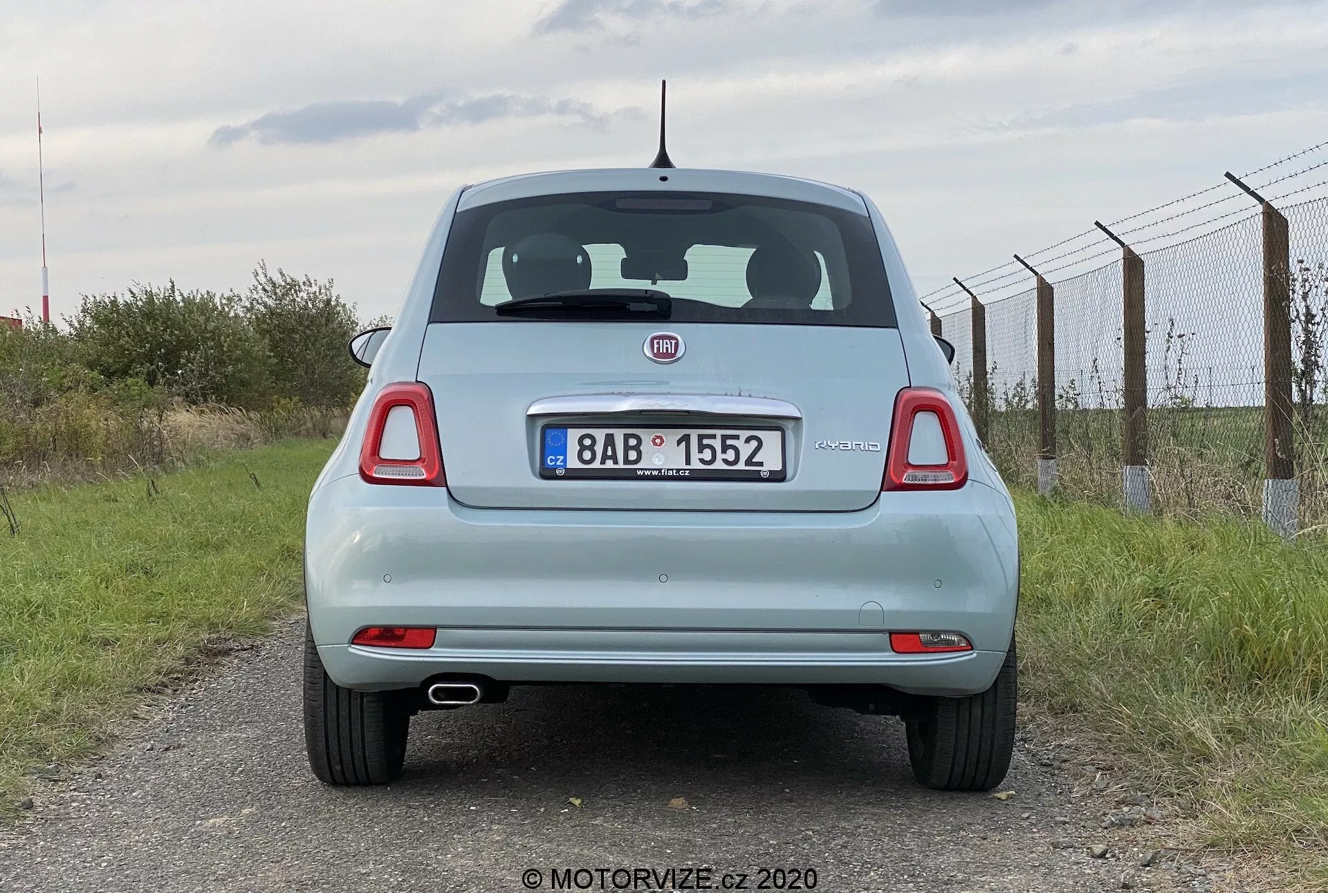 Vista posteriore di una Fiat 500 2015 azzurra. La prospettiva a livello degli occhi cattura il veicolo fermo, mostrando l'intera parte posteriore, compreso il portellone, il lunotto, i fanali posteriori, il paraurti posteriore e la targa. Le caratteristiche includono un emblema del produttore prominente, tergicristallo posteriore, luce di stop alta e fanali posteriori rotondi con segmenti aggiuntivi. La targa è montata al centro del portellone, sotto l'emblema e sopra il paraurti posteriore piatto con riflettori integrati. Un'antenna corta è posizionata nel mezzo del tetto verso la parte posteriore. Cielo nuvoloso e ambiente rurale, con una recinzione a destra e erba intorno.
