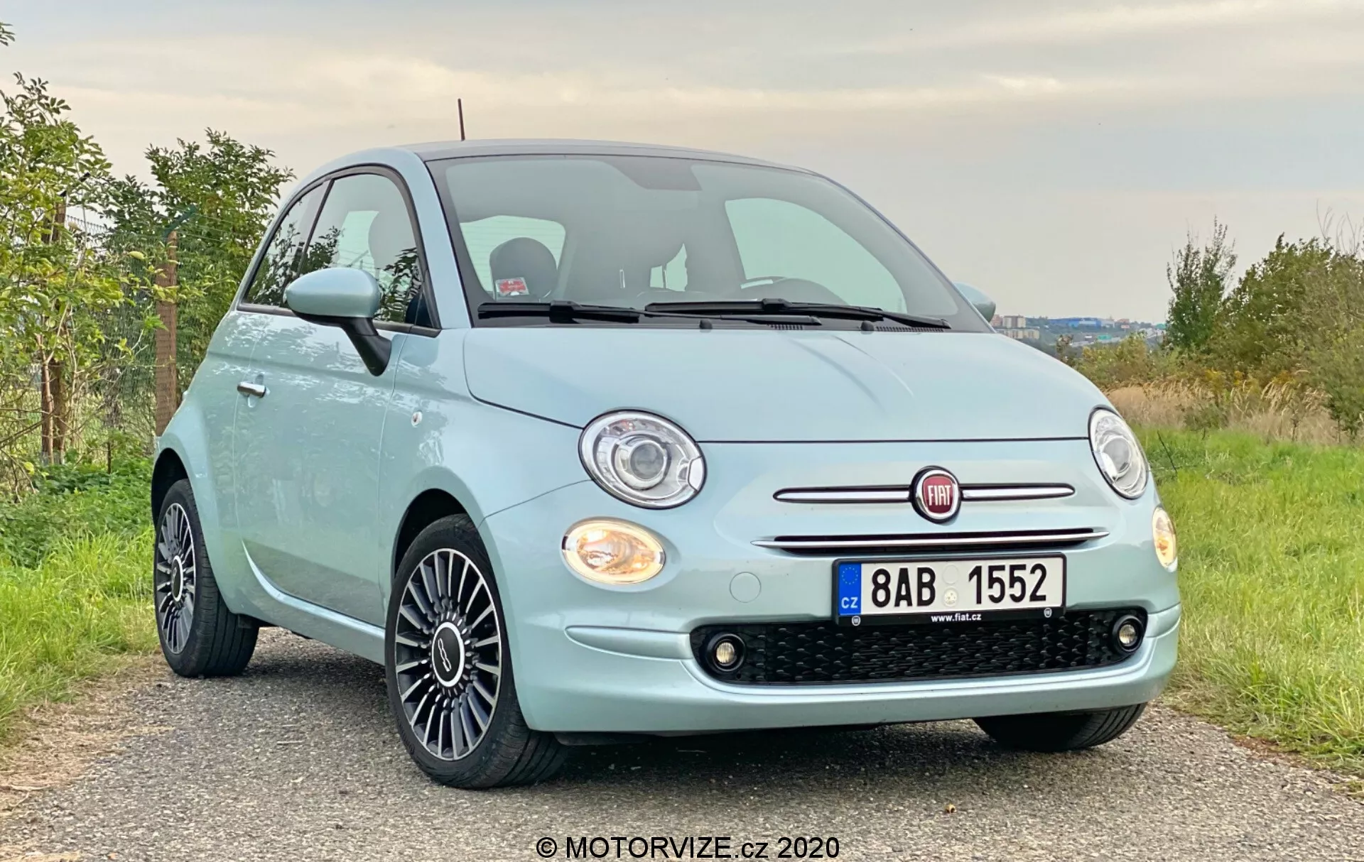 Vista a tre quarti anteriore di una Fiat 500 azzurra del 2015 con griglia cromata e fari alogeni rotondi a un'angolazione leggermente bassa. L'immagine evidenzia la facciata anteriore, la griglia, i fari, il cofano e le ruote in lega con design a raggi multipli neri. L'auto è parcheggiata su una strada sterrata o ghiaiosa in una zona rurale con vegetazione verde sullo sfondo.