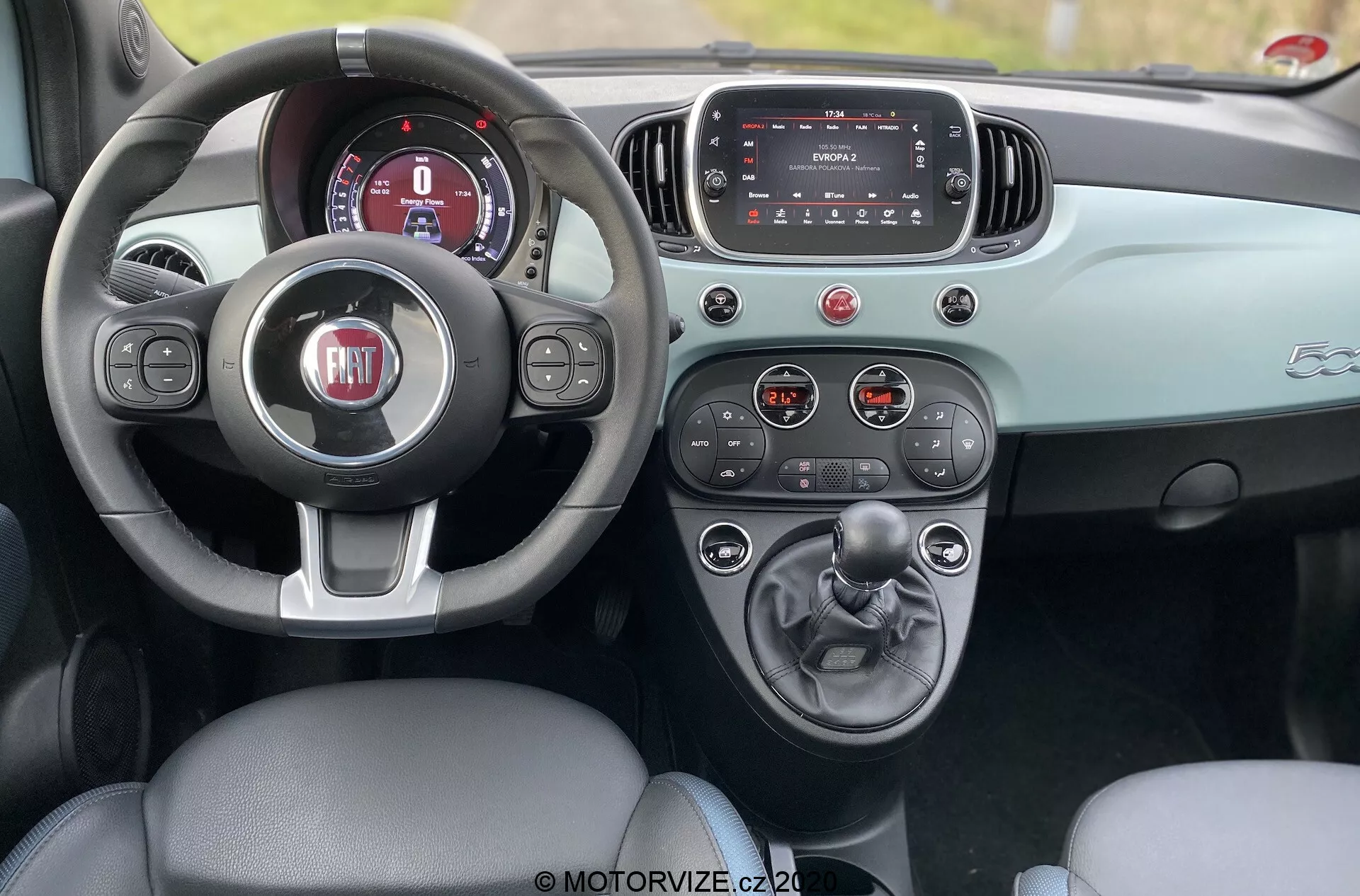 Scatto dell'interno di una Fiat 500 del 2015 dall'area dei sedili posteriori, che mostra il cockpit anteriore con seduta in pelle per il conducente, volante a tre razze con logo FIAT, quadro strumenti digitale, touchscreen dell'infotainment e console centrale con cambio manuale e portabevande. Il cruscotto e la portiera presentano uno schema di colori a due tonalità che esalta il design estetico. La foto è stata scattata durante il giorno, evidenziando il raffinato design degli interni e le funzionalità dell'auto.
