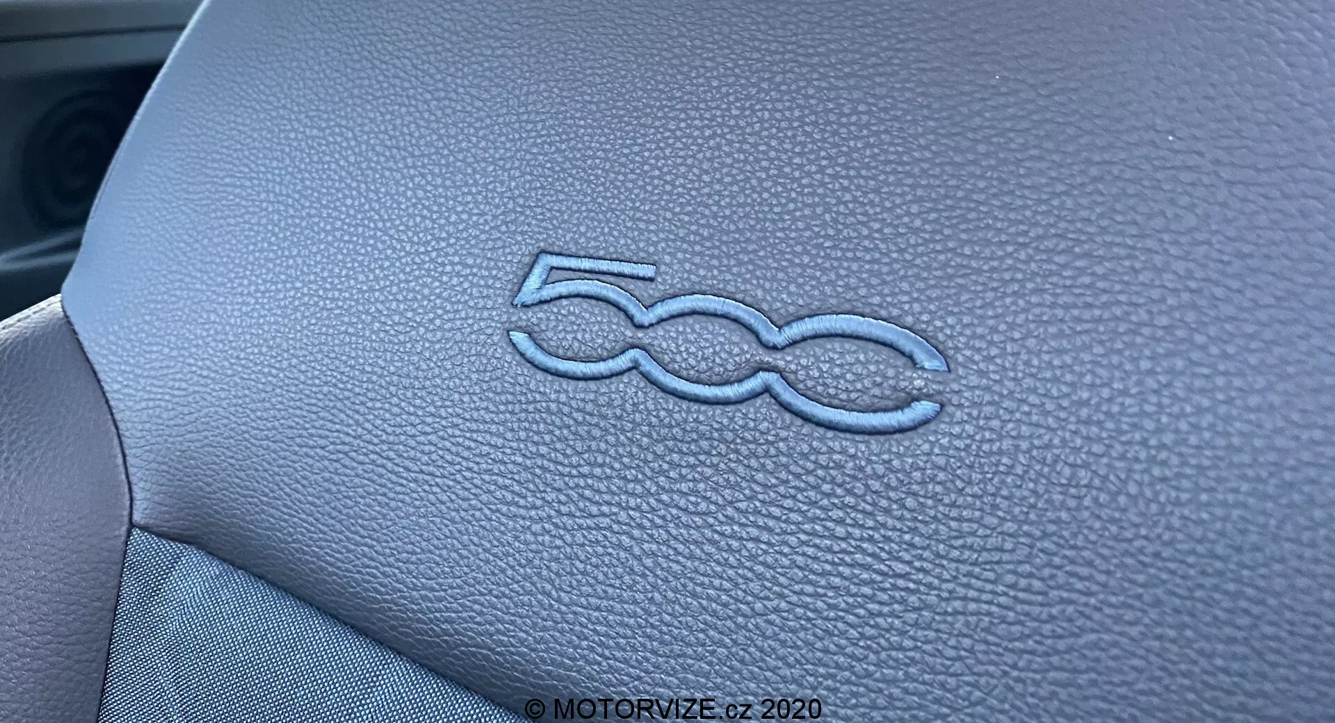 Scatto ravvicinato che mostra il logo '500' stilizzato in rilievo sul cruscotto dell'interno di una Fiat 500 2015. La texture simile alla pelle e le cuciture precise del pannello del cruscotto sono enfatizzate dall'illuminazione e dalla ridotta profondità di campo, mettendo in evidenza la qualità artigianale del design degli interni del veicolo.