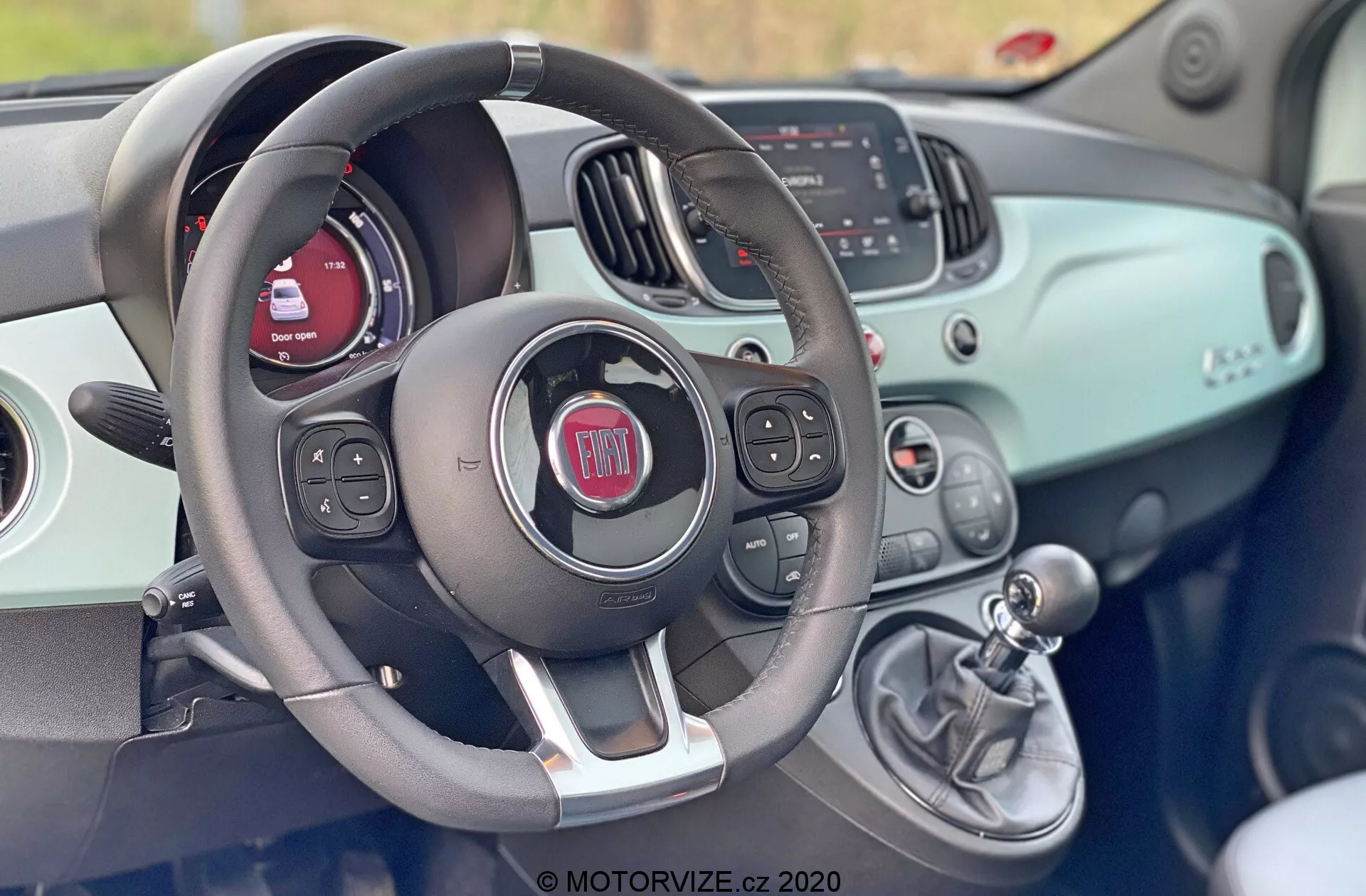 Vista interna di una Fiat 500 del 2015 dal sedile del conducente che mostra il volante con comandi integrati per l'audio, i comandi vocali e forse il cruise control. Dietro al volante, il cruscotto presenta uno schermo digitale e indicatori tradizionali. Il cruscotto ha una superficie dello stesso colore della carrozzeria e include un display per l'infotainment che mostra informazioni su radio e media, oltre a comandi HVAC manuali. A destra del volante, una leva del cambio manuale indica una trasmissione manuale. Il design complessivo è moderno con un'estetica sportiva e bicolore.