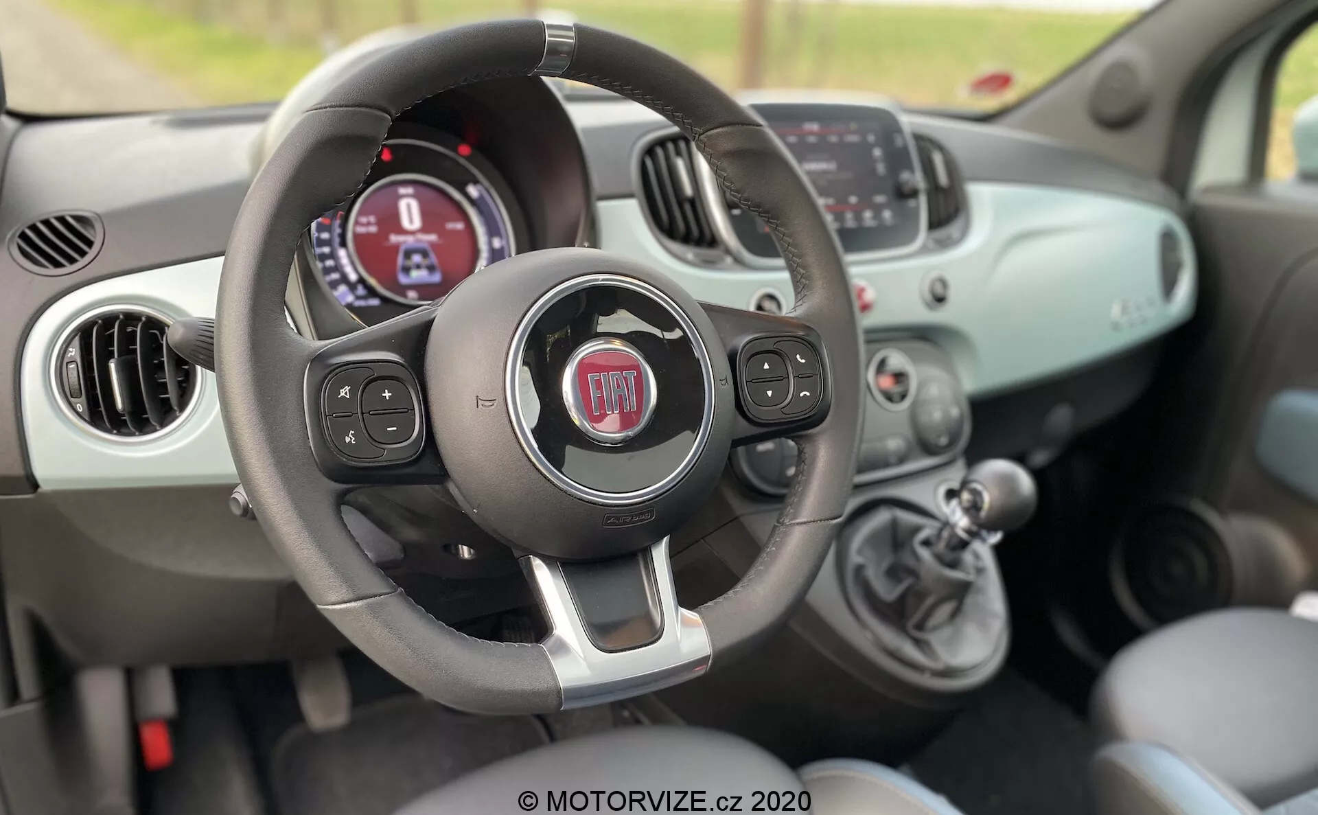 Vista interna di una Fiat 500 del 2015 dal lato passeggero, che mostra il volante con pulsanti multifunzione, il cruscotto con display digitale, i comandi del clima manuali e la leva del cambio.