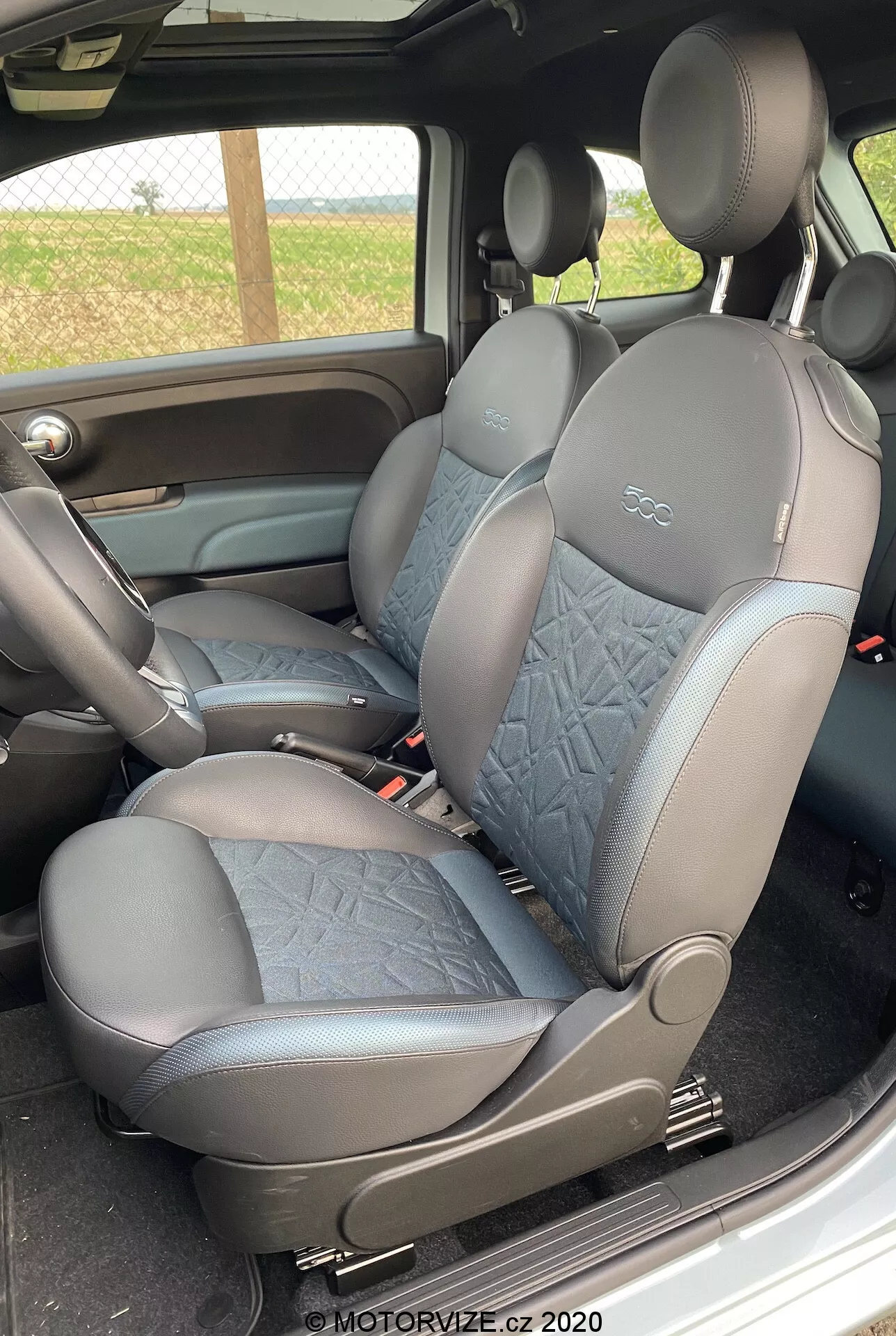 L'immagine mostra l'interno di una Fiat 500 2015 nell'area passeggeri, sedili anteriori da angolazione laterale. Rivestiti in pelle e tessuto trapuntato. Poggiatesta integrati. Tonalità scura per praticità. Lev lever di regolazione manuale del sedile visibile. Console centrale con bracciolo parzialmente visibile. Coerenza cromatica e dei materiali con il pannello porta. Filigrana del 2020.