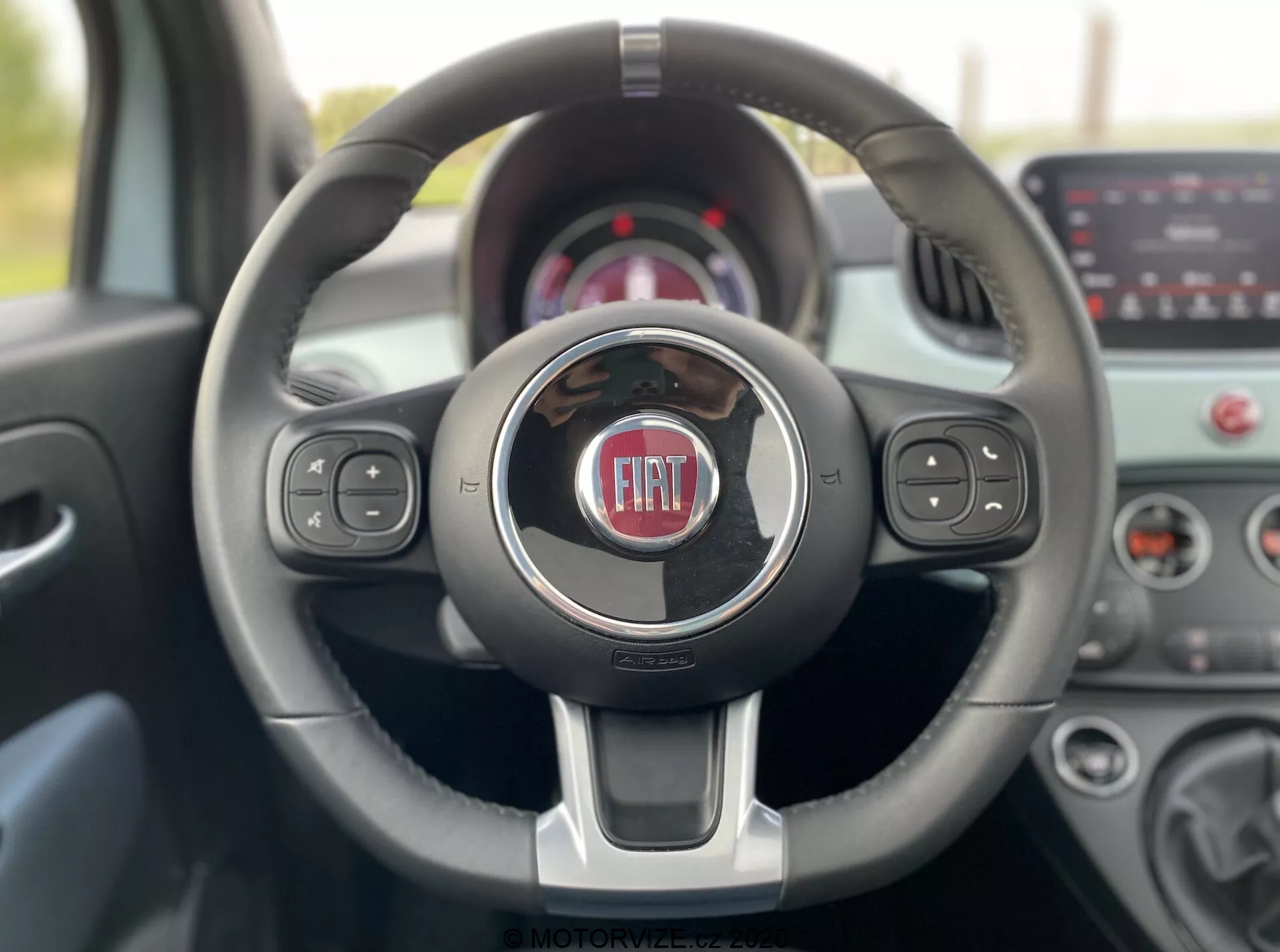 Vista interna di una Fiat 500 2015 dal posto di guida, che mostra un volante a tre razze moderno con accenti cromati e il logo Fiat al centro. Il volante include comandi integrati per media, funzioni vocali e controllo della velocità. Lo sfondo sfocato presenta il quadro strumenti e il sistema di infotainment touchscreen, evidenziando la tecnologia e il design contemporanei del veicolo.