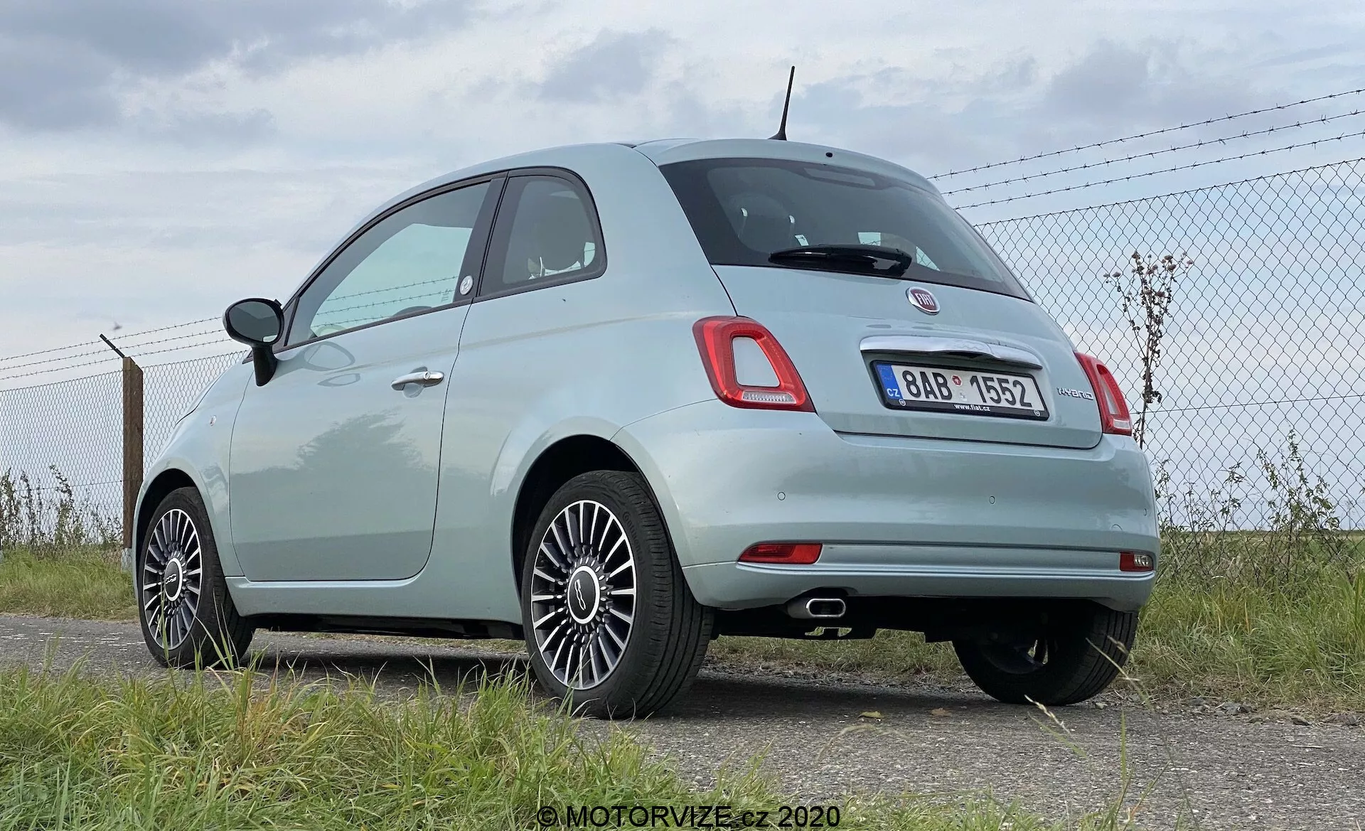Vista posteriore tre quarti di una moderna Fiat 500 hatchback di colore chiaro, che mostra i caratteristici fanali posteriori arrotondati e un badge cromato centrale sopra l'area della targa posteriore. L'immagine mette in evidenza la linea del tetto arrotondata del veicolo, le modanature cromate intorno ai finestrini laterali e i cerchi in lega multirazza. La vernice pastello chiara dell'auto indica che è relativamente nuova o ben mantenuta. È parcheggiata vicino a una recinzione a catena su un'area asfaltata, con il cielo e l'orizzonte visibili sullo sfondo, suggerendo un campo d'aviazione o uno spazio aperto. Le possibili caratteristiche tecnologiche includono sensori di parcheggio posteriori integrati nella sezione nera del paraurti posteriore.