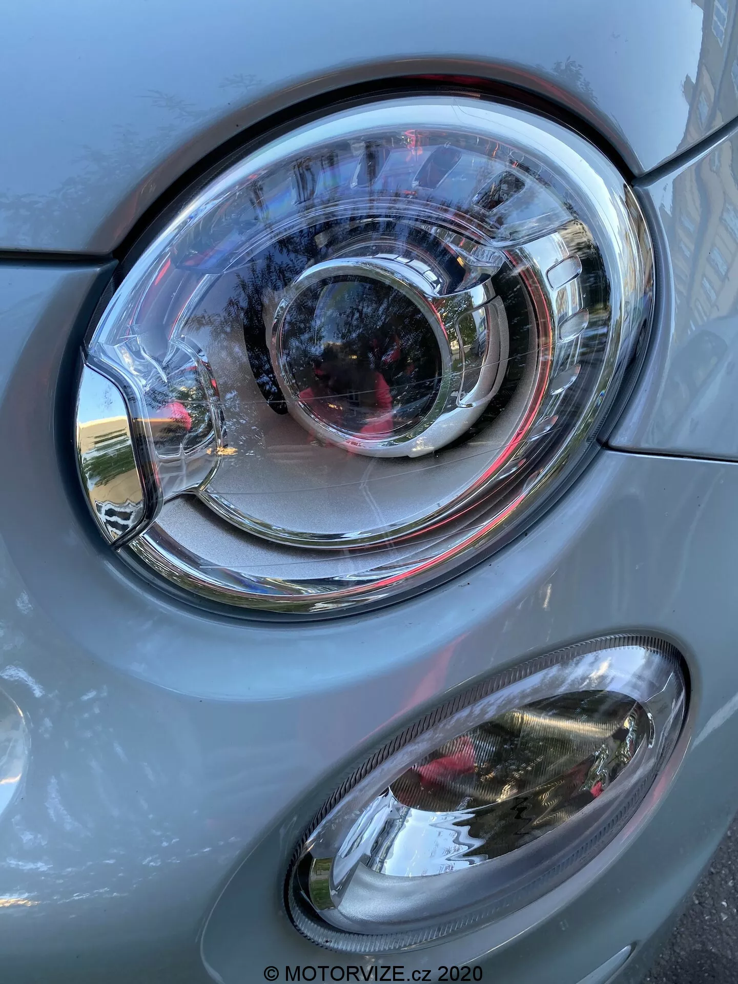 Vista ravvicinata del lato anteriore sinistro di una Fiat 500 del 2015, con un focus sull'assieme del faro. L'immagine, in orientamento verticale, mostra un prominente faro principale con lente in policarbonato chiara dal design complesso di anelli concentrici, che suggeriscono uno stile a proiettore e possibilmente tecnologia di illuminazione adattiva. Sotto il faro principale si trova una luce secondaria antinebbia di forma ovale con una copertura chiara. I pannelli della carrozzeria dell'auto, di colore grigio con riflessi metallici, hanno una finitura liscia che riflette la luce del giorno e elementi dell'ambiente circostante come edifici e cielo. Il gruppo ottico e la luce antinebbia sono in buone condizioni, a indicare un veicolo ben tenuto o nuovo.