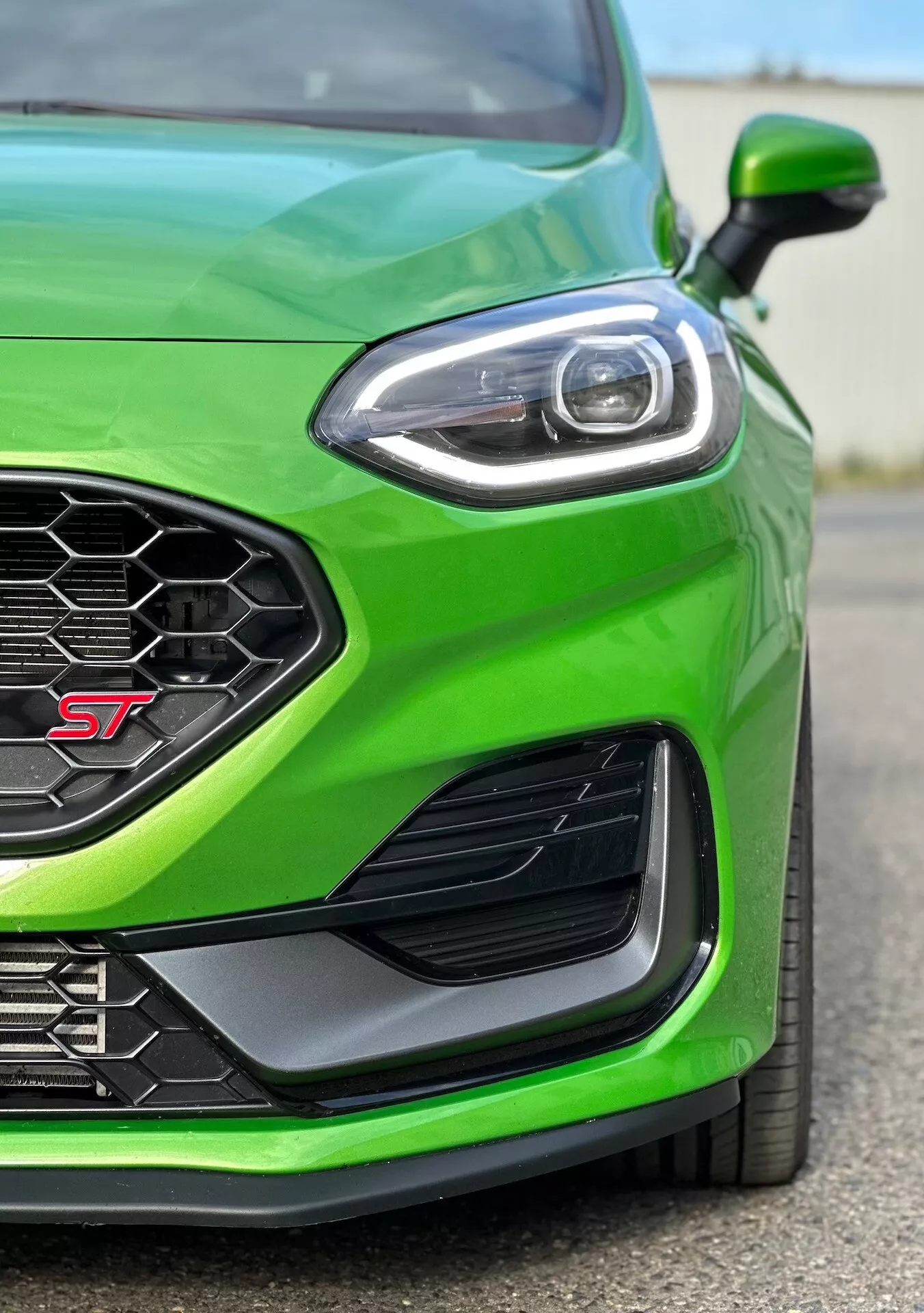 Vista ravvicinata del quarto anteriore sinistro di una Ford Fiesta ST 2022 in verde brillante, mettendo in evidenza il trim orientato alle prestazioni, griglia a nido d'ape per flusso d'aria ottimale, design avanzato dei fari LED, presa d'aria laterale aerodinamica e paraurti anteriore per una migliore canalizzazione dell'aria.