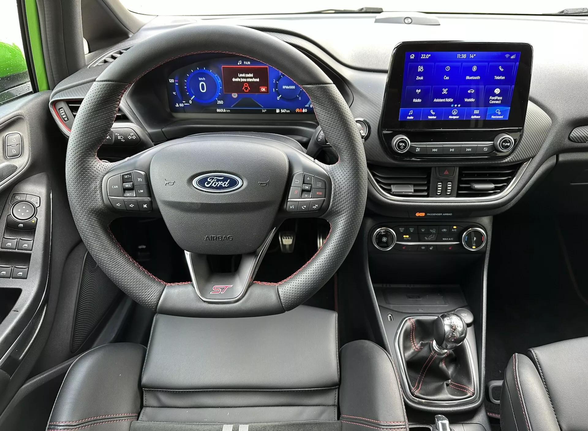 Vista interna della Ford Fiesta 2022 dal sedile posteriore, evidenziando il volante con logo Ford e badge ST, pannello strumenti digitale, sistema di infotainment touchscreen, leva del cambio manuale e sedile del conducente sportivo con cuciture a contrasto.
