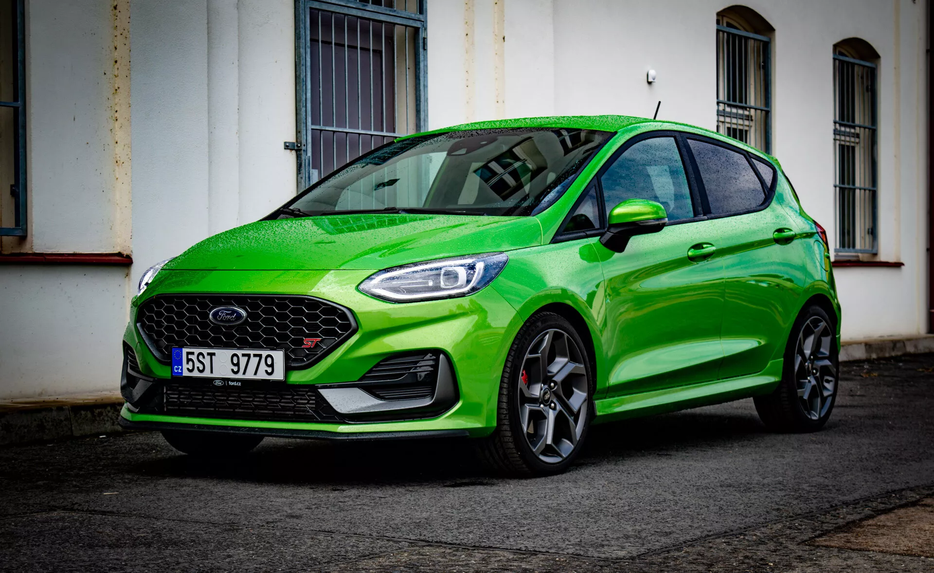 Ford Fiesta 2022 in verde brillante, che mostra il suo design sportivo con una vista angolare bassa frontale tre quarti che evidenzia il paraurti anteriore, la griglia, i fari, il cofano, il parafango, le ruote e il riflesso del parabrezza su una strada urbana dopo la pioggia