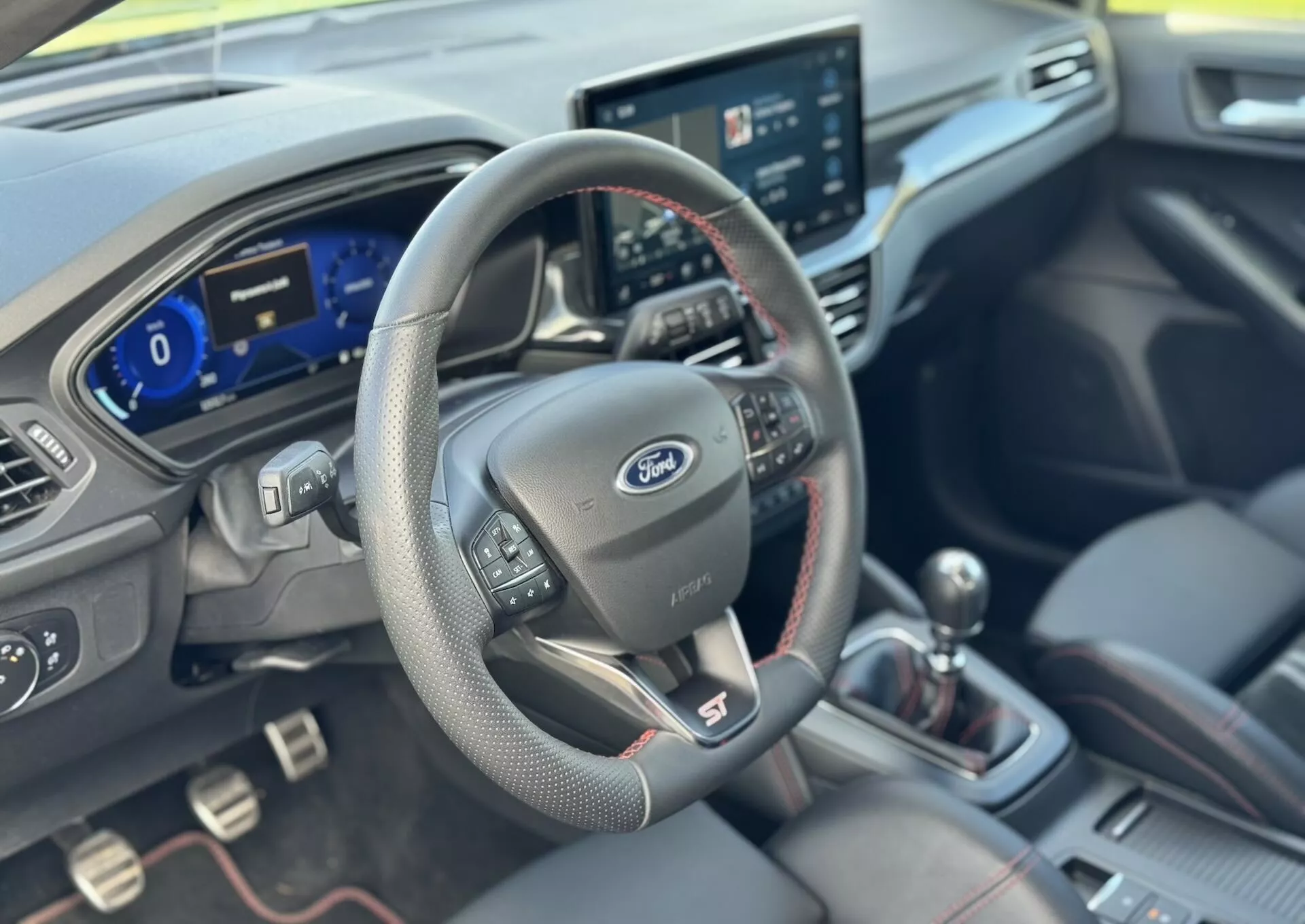 Interni Ford Focus ST 2022 che mostrano la prospettiva del conducente con volante rivestito in pelle dotato di controllo multimediale e cruise control, cruscotto digitale, sistema di infotainment centrale touchscreen, leva del cambio manuale con cuciture rosse e sedili premium imbottiti