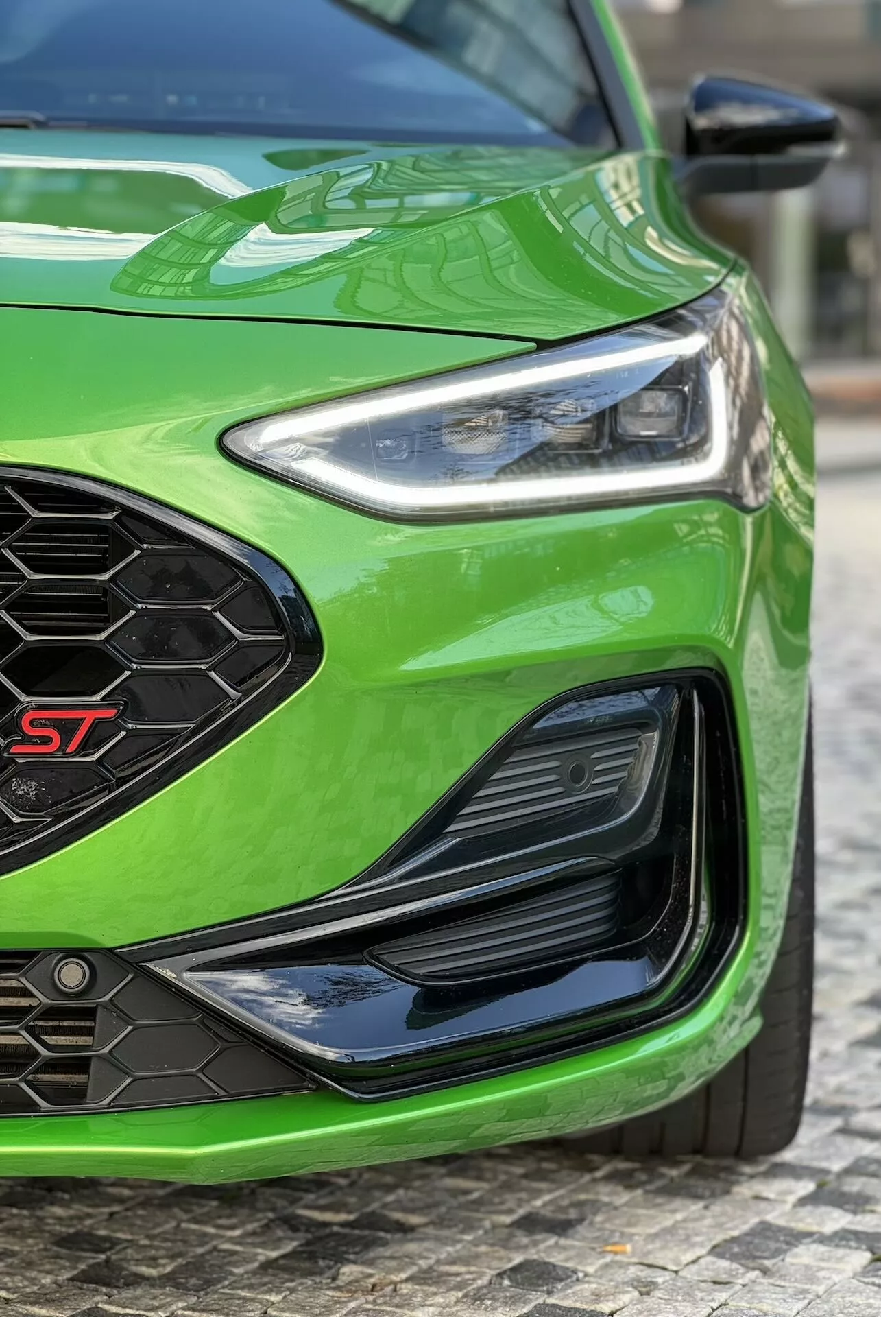 Vista angolare e dettagliata della sezione anteriore sinistra di una Ford Focus ST 2022 in verde brillante, che mostra il faro LED, la griglia frontale a nido d'ape con badge ST, il paraurti inferiore con prese d'aria e la finitura della vernice lucida, riflettendo funzionalità avanzate di illuminazione, aerodinamica e prestazioni.