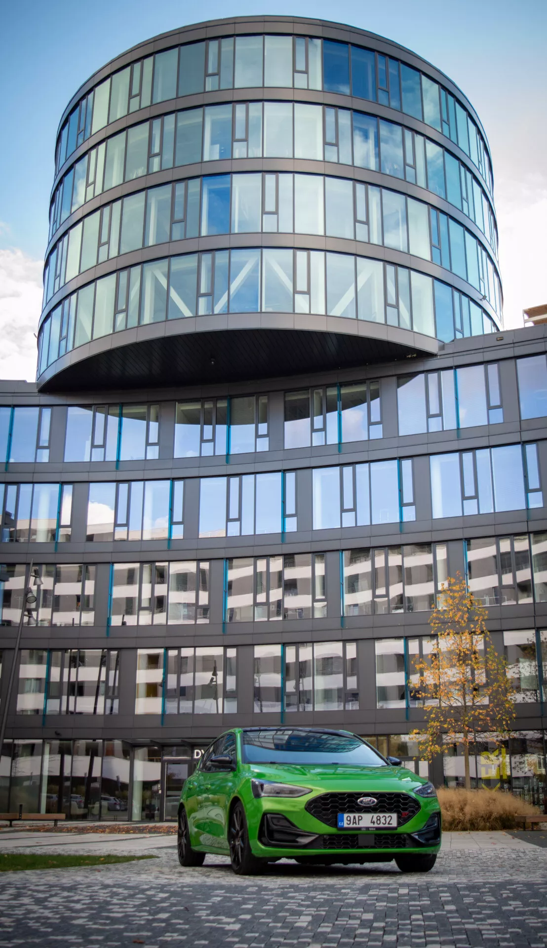 Ford Focus ST 2022 in verde vivace posizionata all'aperto con uno sfondo di edificio in vetro che mette in mostra i suoi fari eleganti, griglia frontale prominente, ruote in lega e design aerodinamico